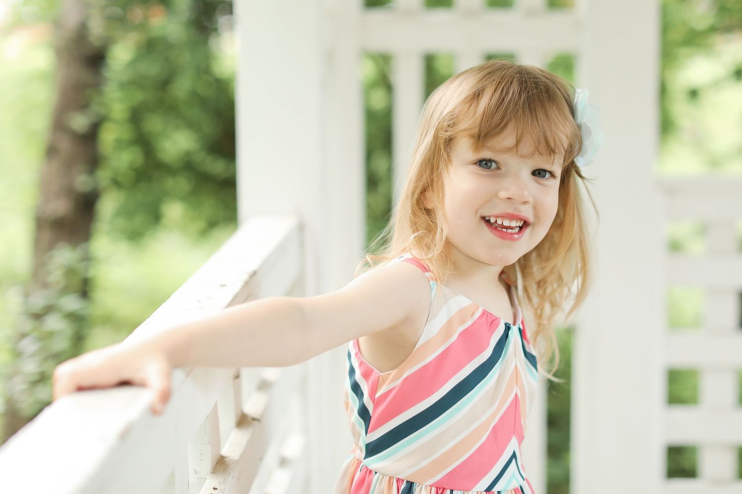 Child in striped dress twirling on white porch in summer light.
