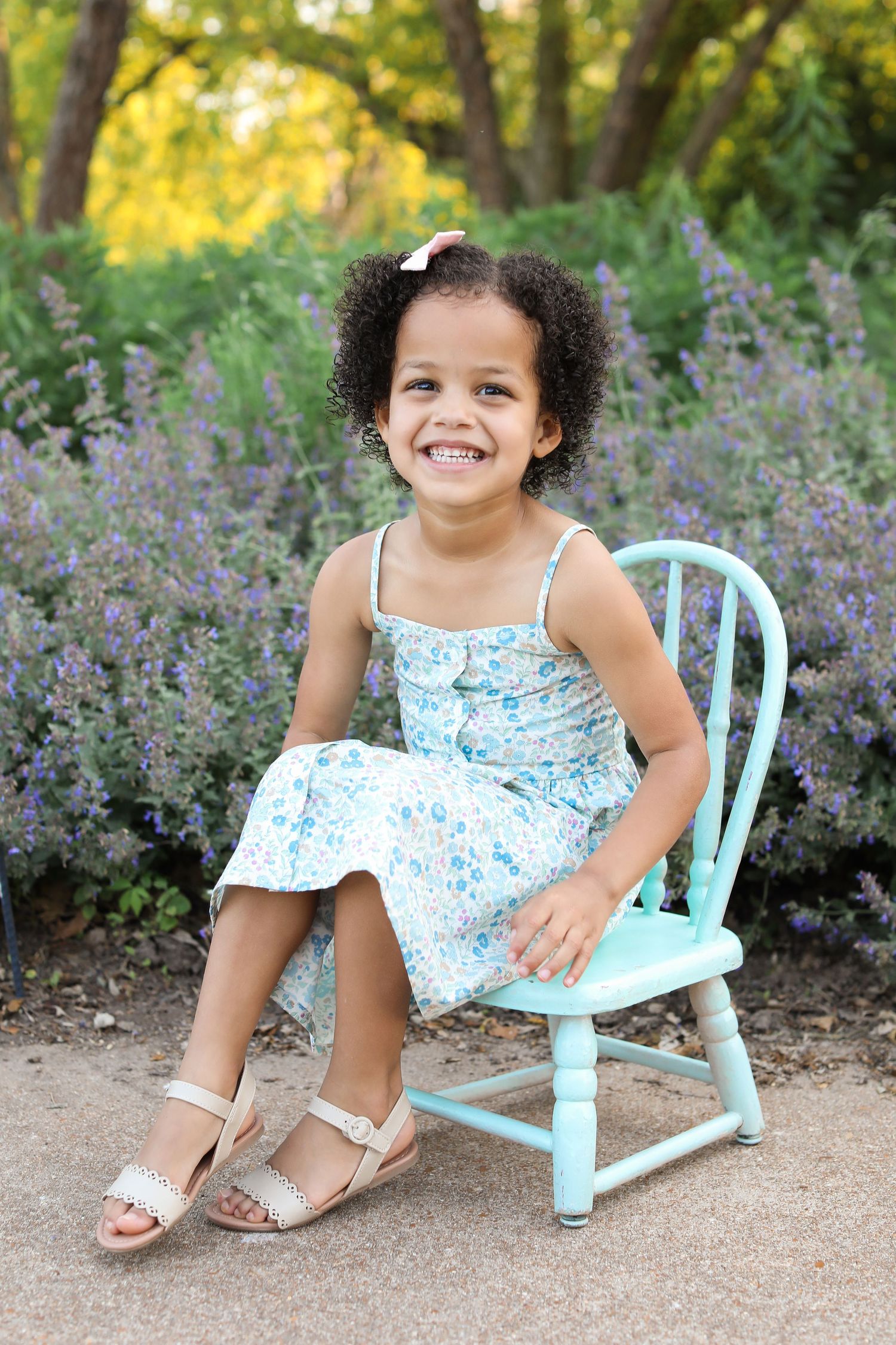 Child in floral dress sitting on light blue wooden chair among purple flowers.