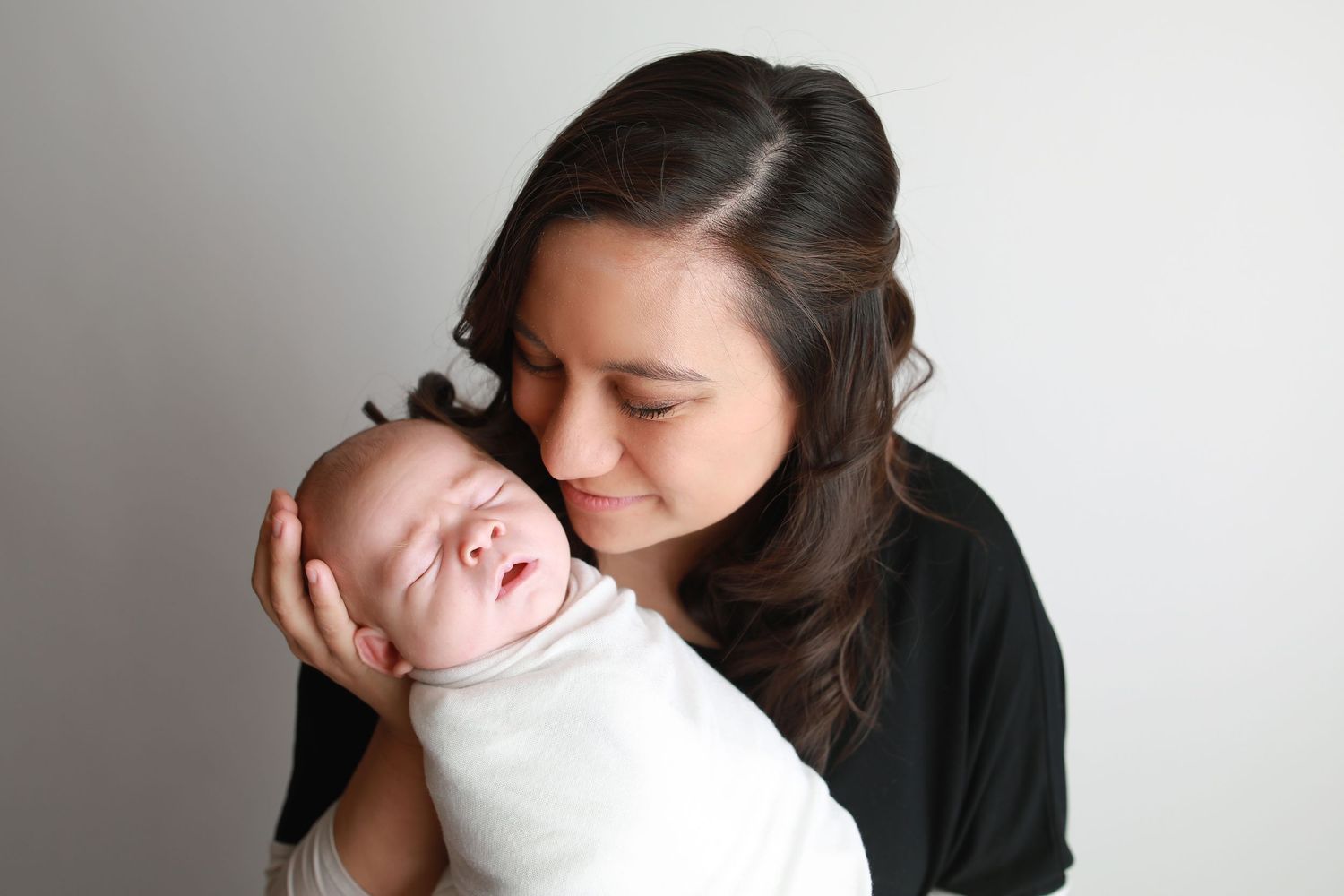 Woman in black cuddles sleeping newborn wrapped in white blanket against white background.
