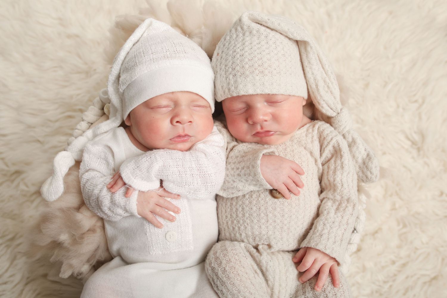 Twin newborns in white and gray knit outfits sleeping side by side.
