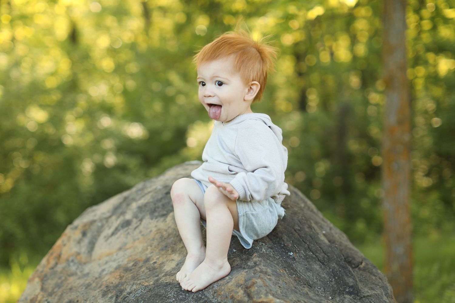 Small child in white outfit sitting on large boulder in sunlit forest setting.