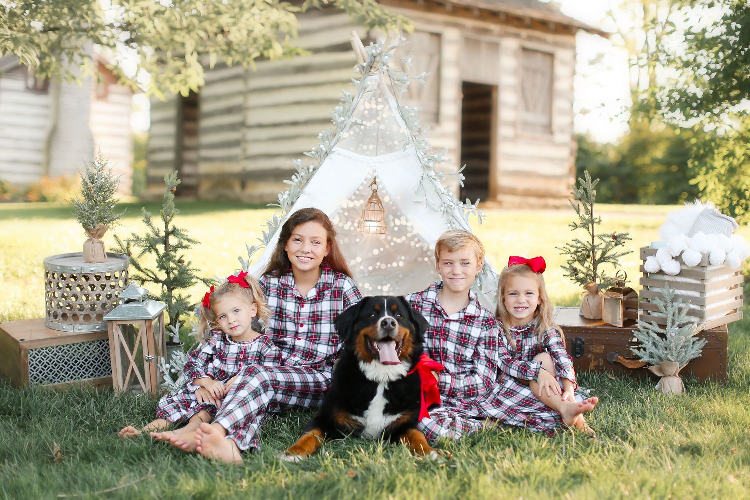 Holiday portrait of matching plaid-dressed group with dog in front of decorated teepee and rustic cabin.
