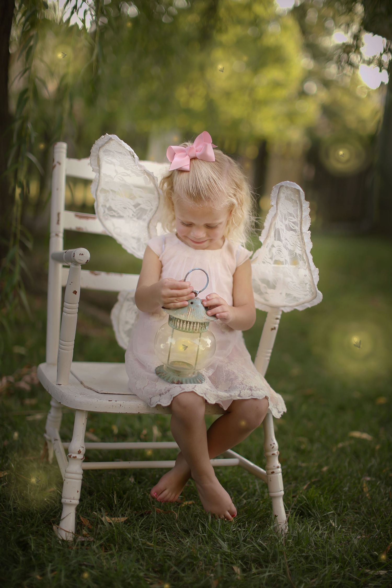 Small figure in white dress with wings holds lantern while sitting on antique white chair in dreamy garden setting.