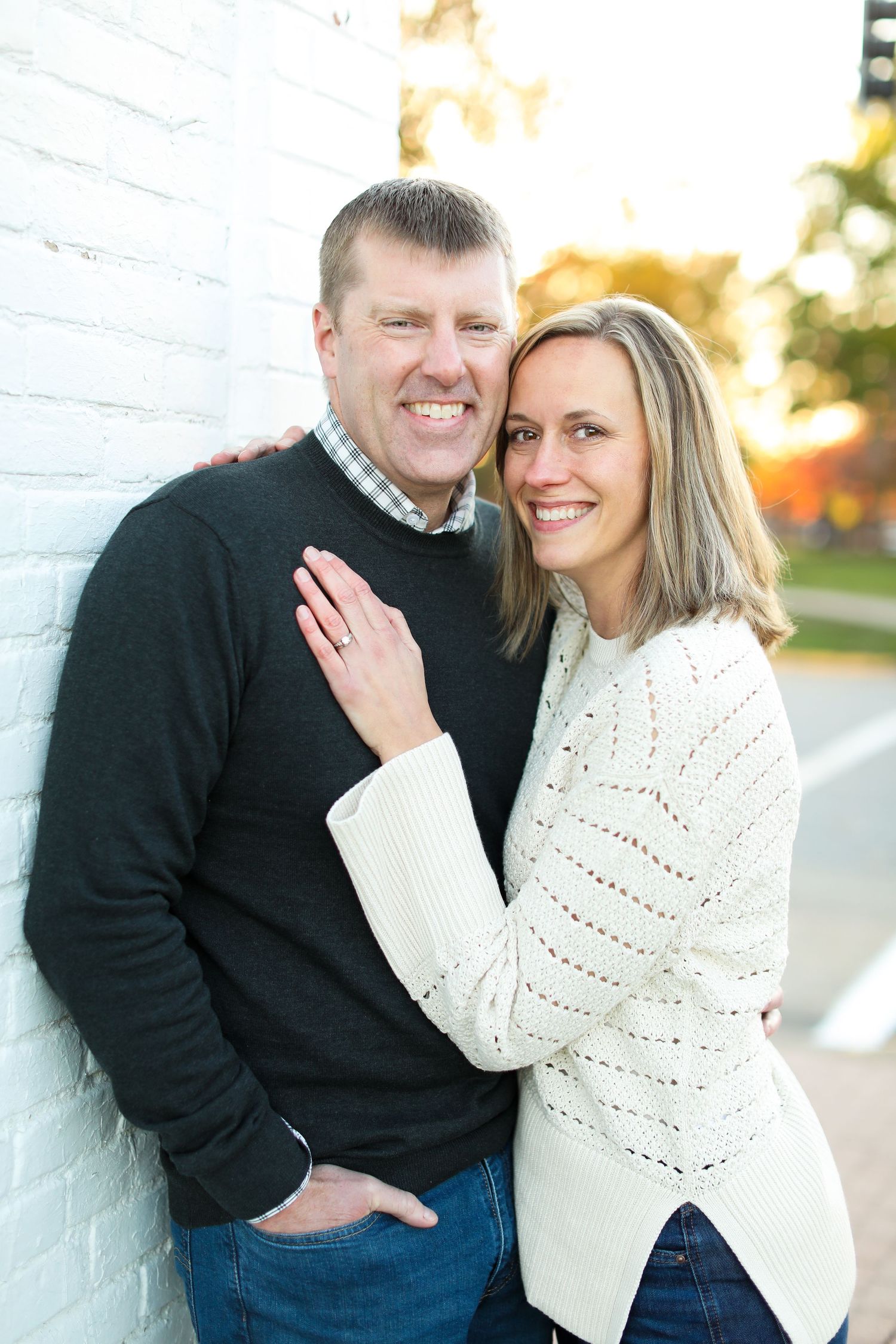 A couple poses against a white wall outdoors during autumn.