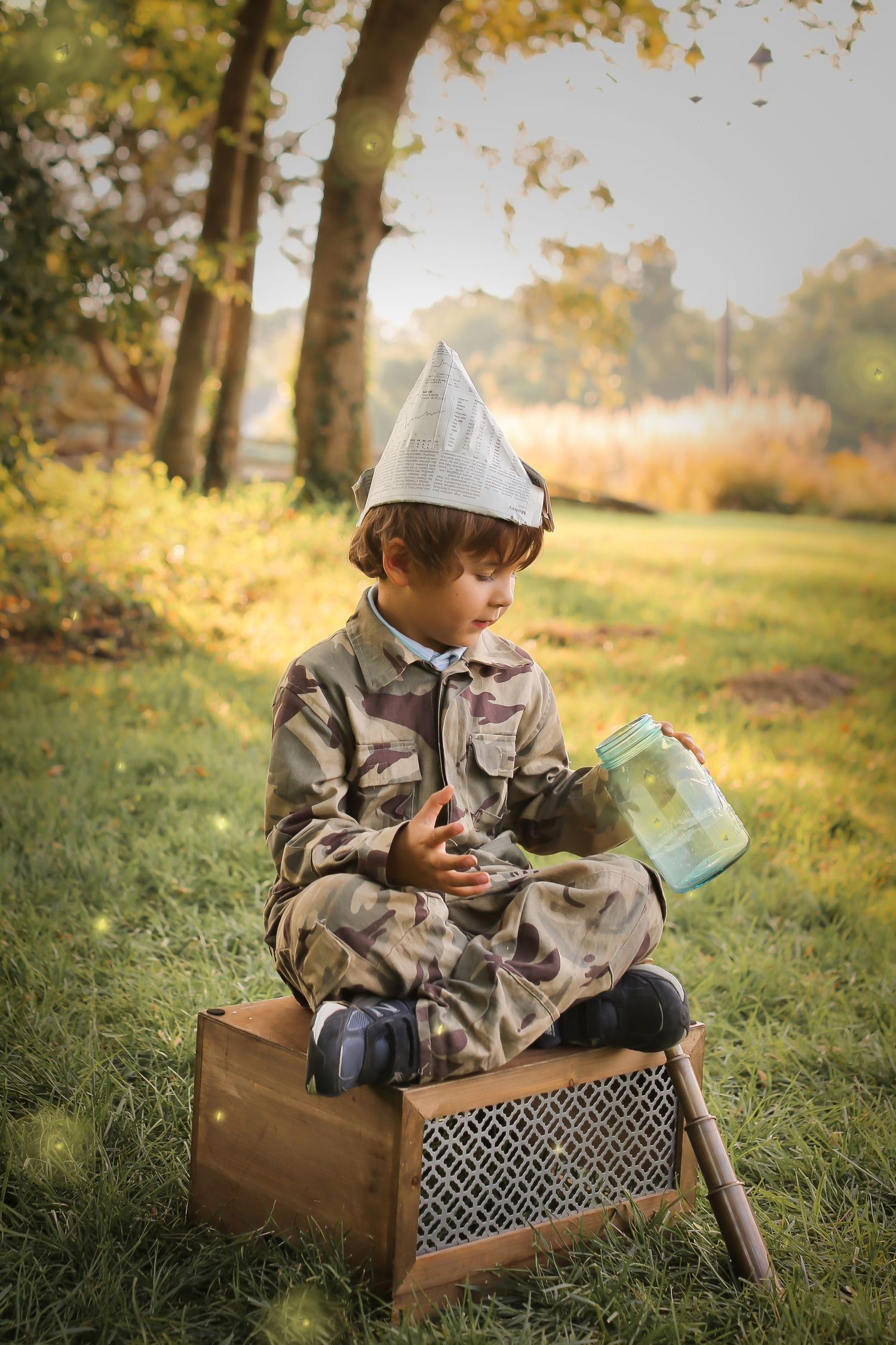 Young adventurer in camo and tin foil hat reads map while sitting on vintage box in autumn sunlight.