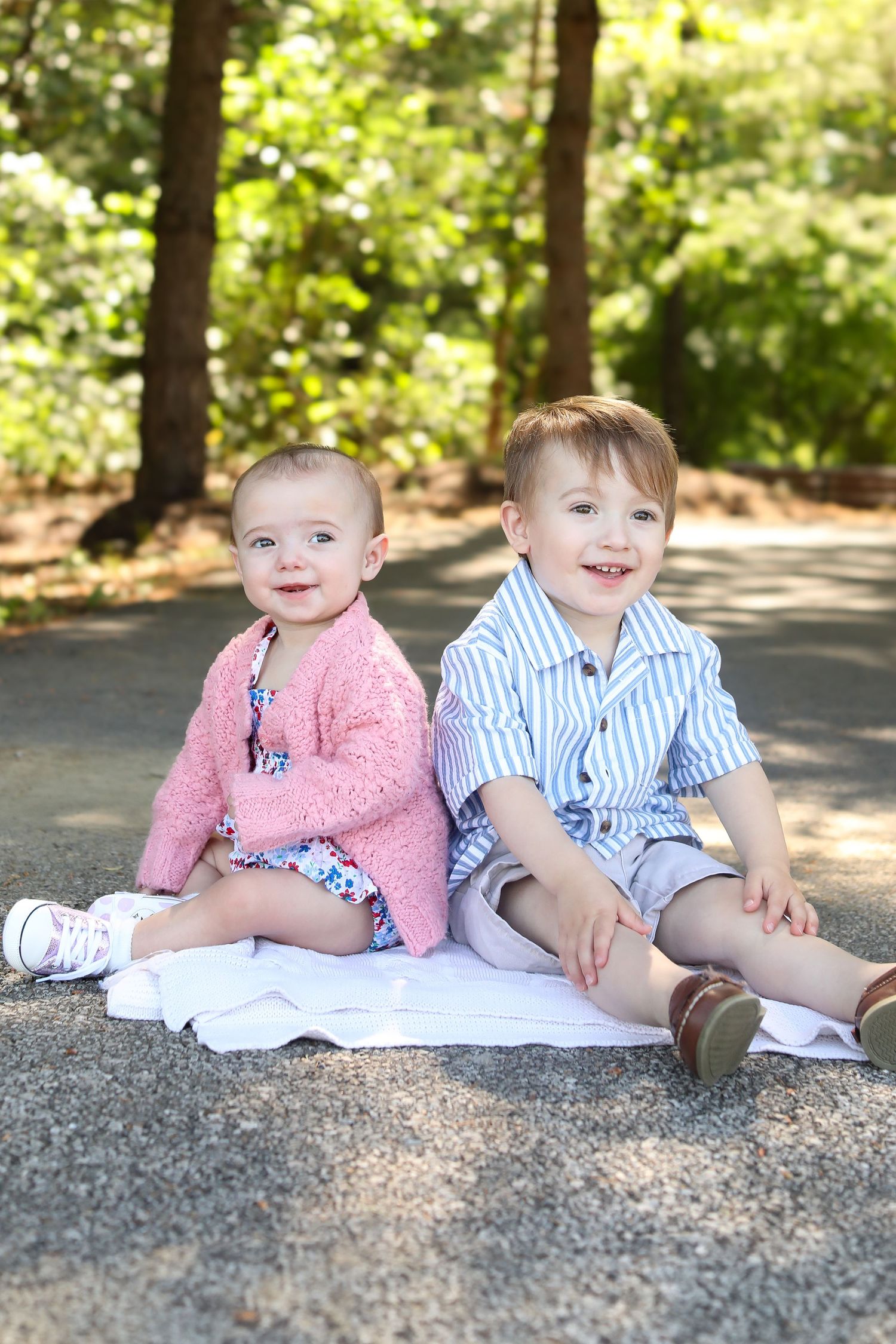 Two young children sitting together on a blanket in a shaded outdoor setting.