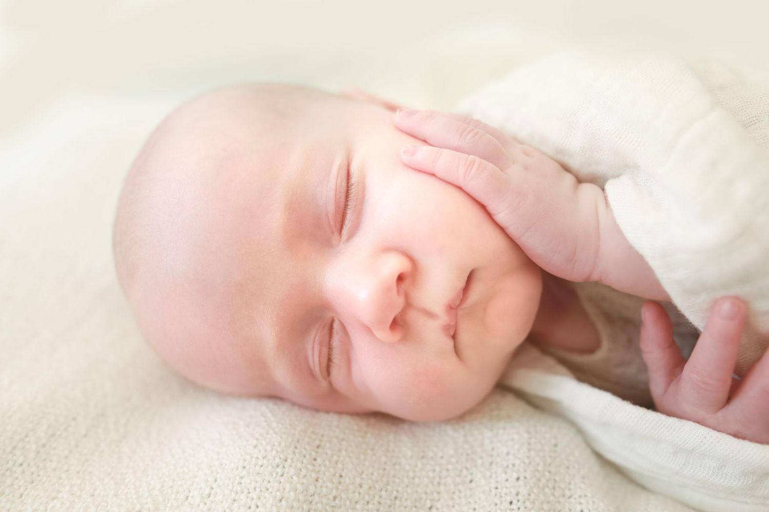 Peaceful sleeping newborn baby with tiny hand resting on soft cream fabric.