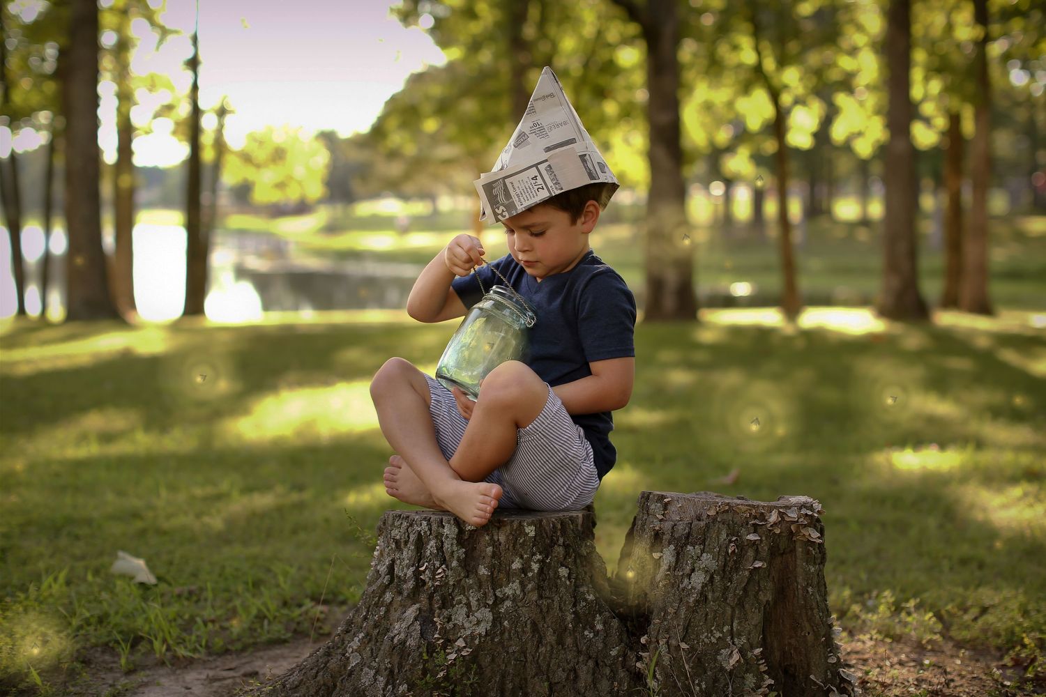 Child sitting on tree stump wearing newspaper hat in sunlit forest.