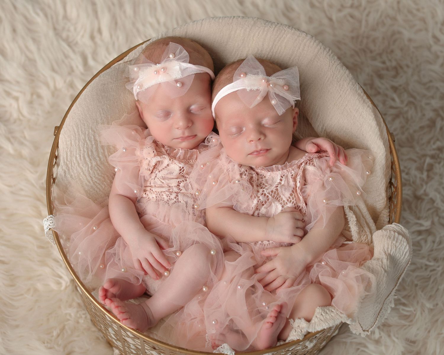 Twin baby girls in pink lace dresses sleeping in round basket on cream fur.