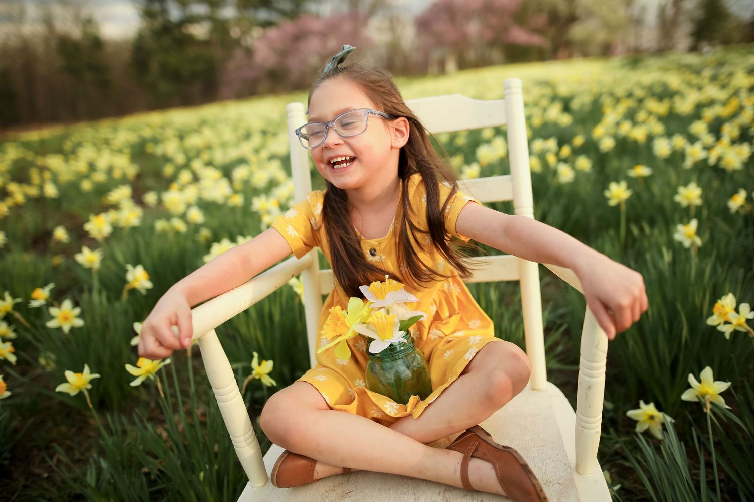 Child in yellow dress laughing joyfully in white chair surrounded by daffodils.