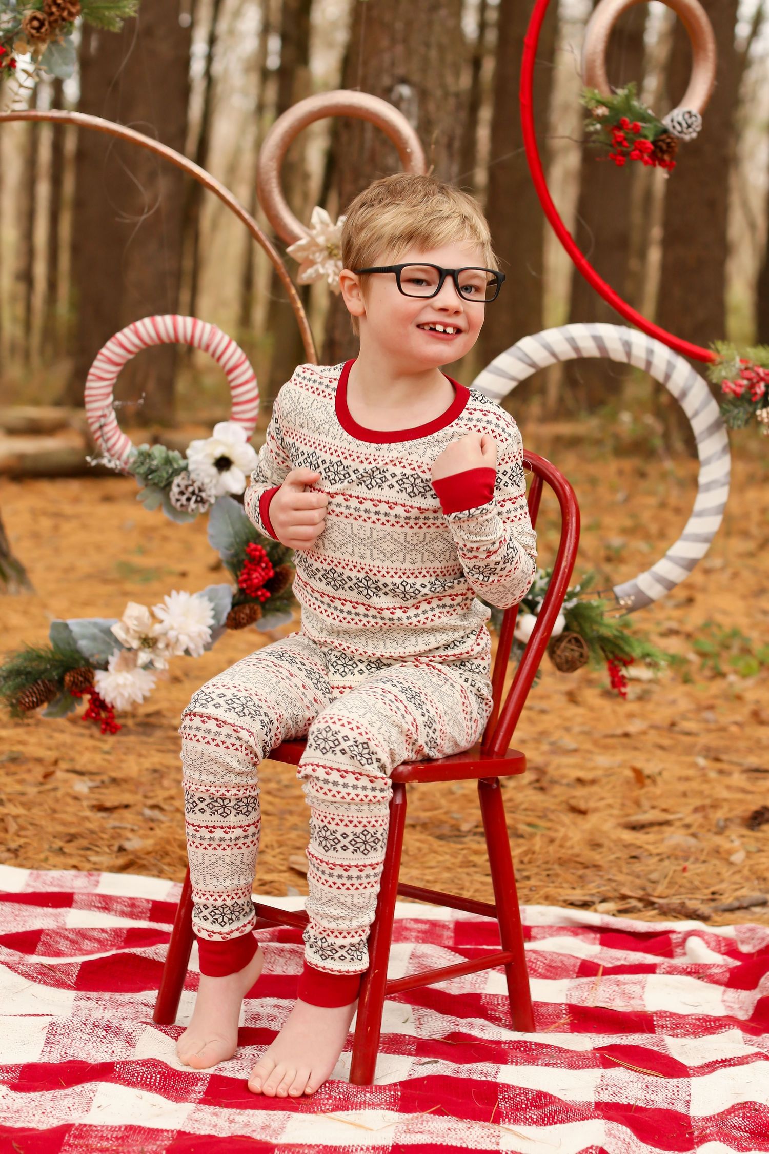 Winter themed photo shoot with red chair and checkered blanket on forest floor with holiday decorations.