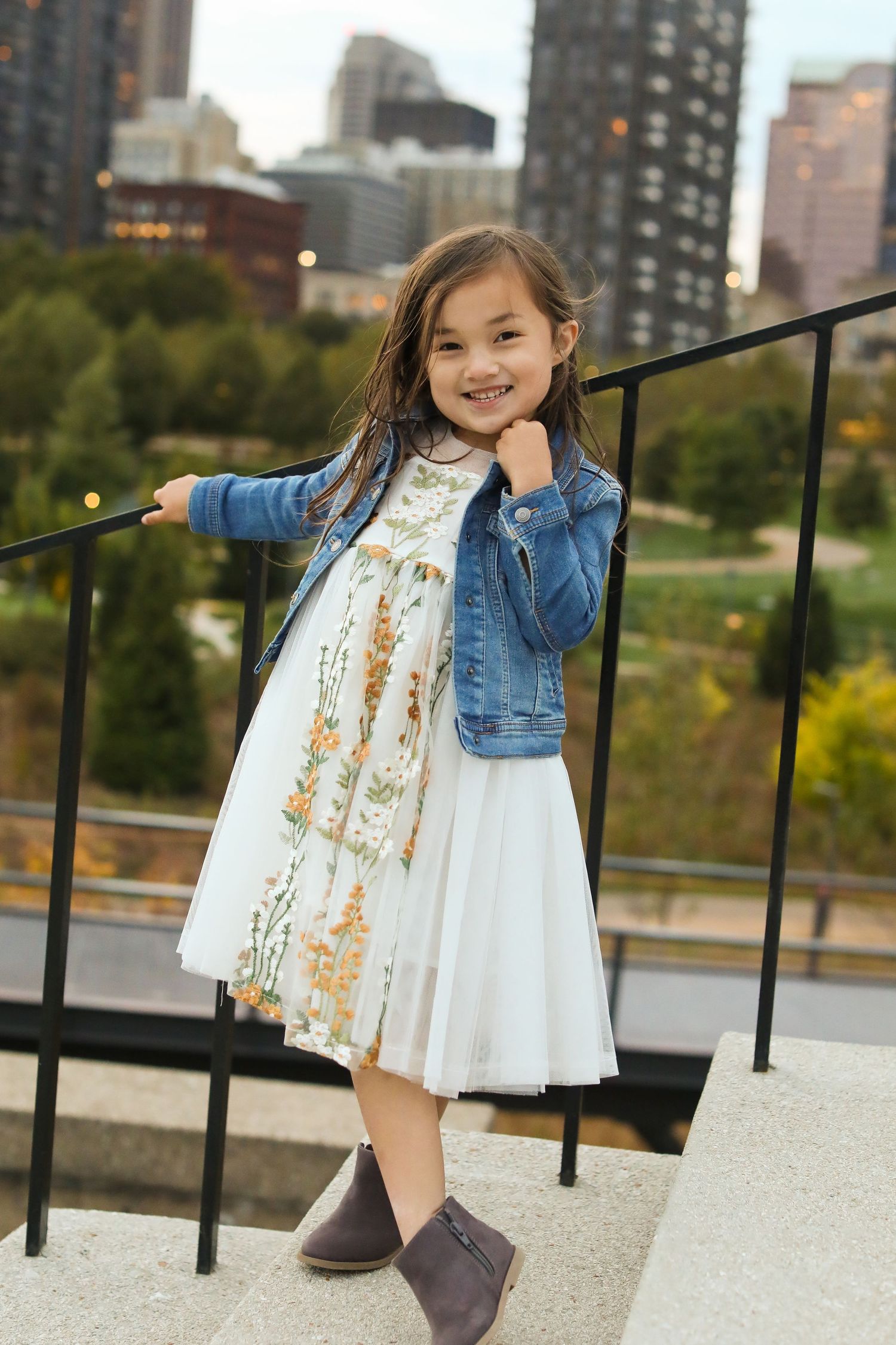 Young girl in white dress and denim jacket poses playfully near urban railing.