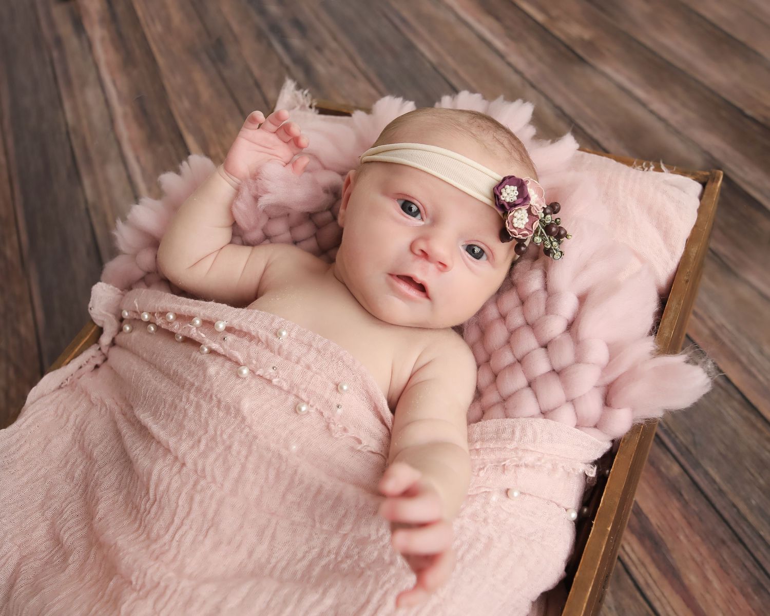 Baby in pink headband lying on tufted pink cushion against wooden backdrop.