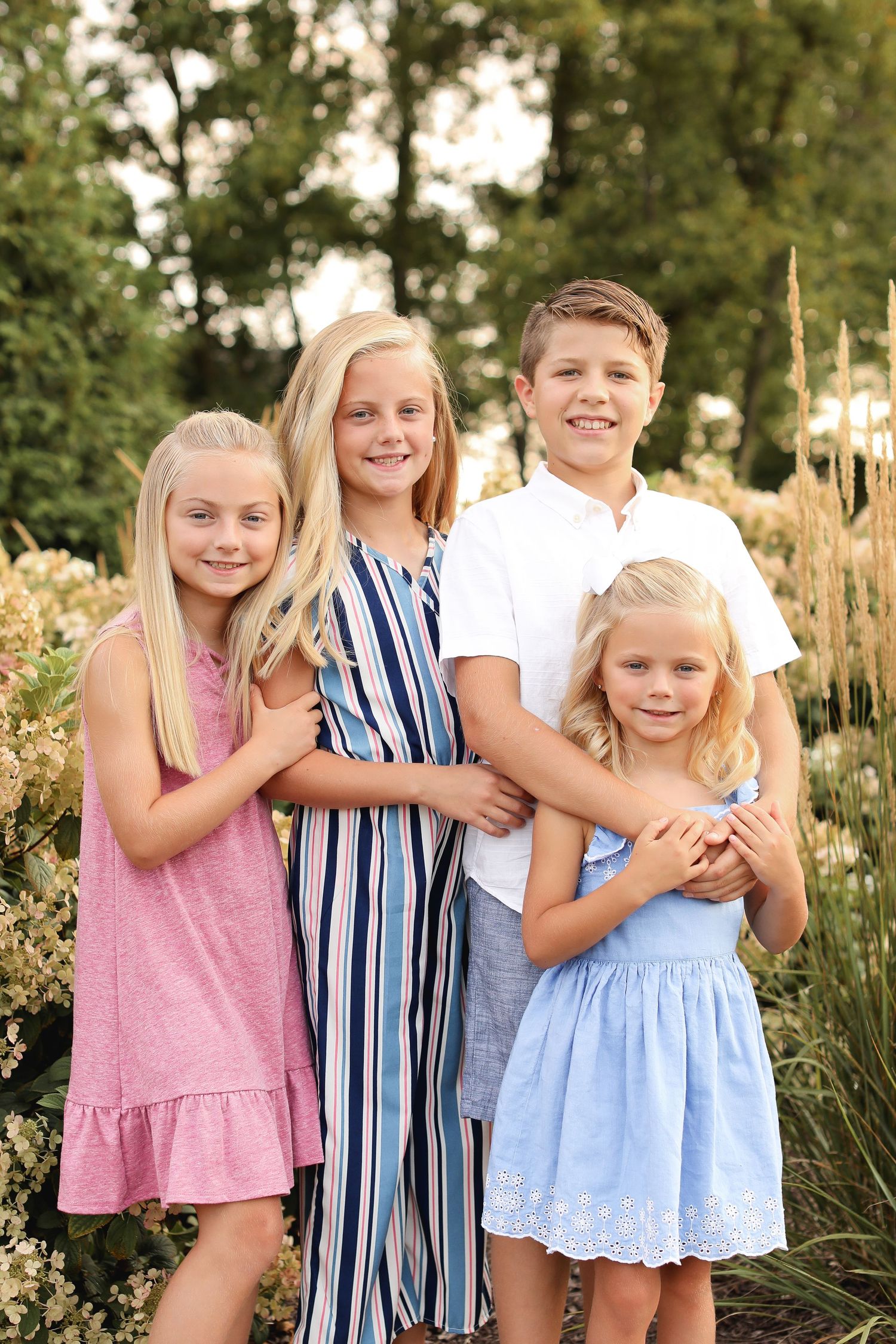 Four children in summer dresses and casual wear pose together in a garden setting.