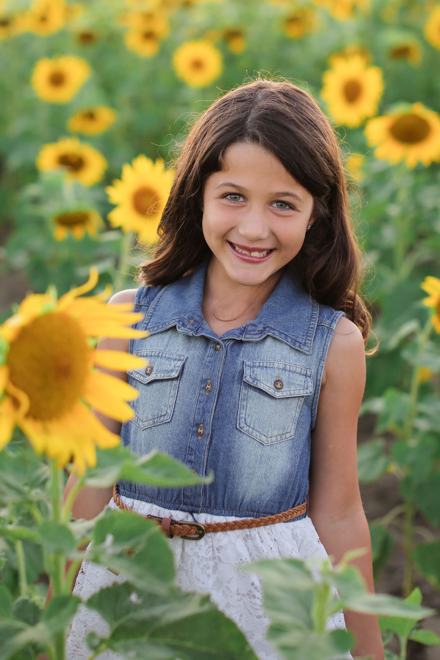 Denim vest-clad figure poses with bright sunflowers in blooming summer garden.