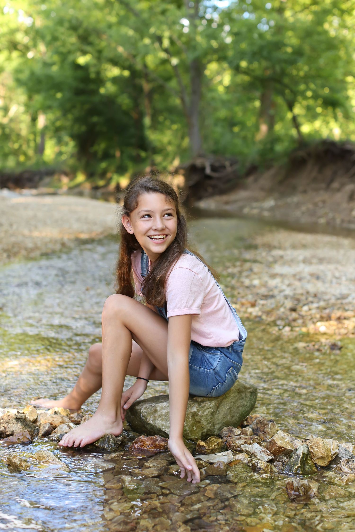 Young person sitting barefoot on rocks by a creek in summer wearing jean shorts and pink shirt.