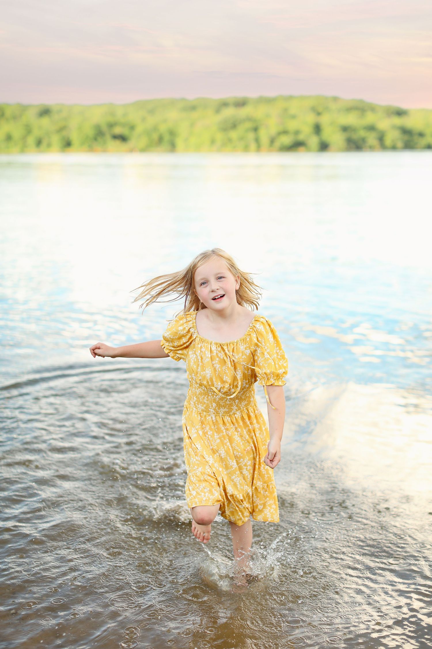 Child splashing in lake water wearing yellow summer dress at sunset.