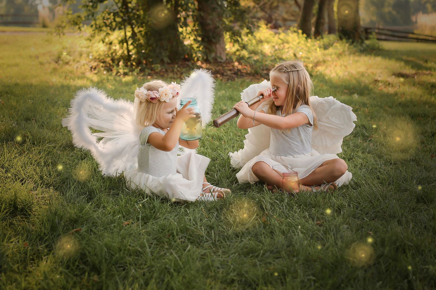 Two fairy-wing adorned figures sit on grass in dreamy golden sunlight during a magical outdoor photoshoot.