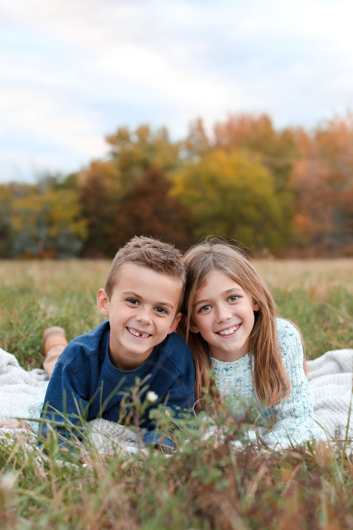 Two children lying on a blanket in a field during fall season.