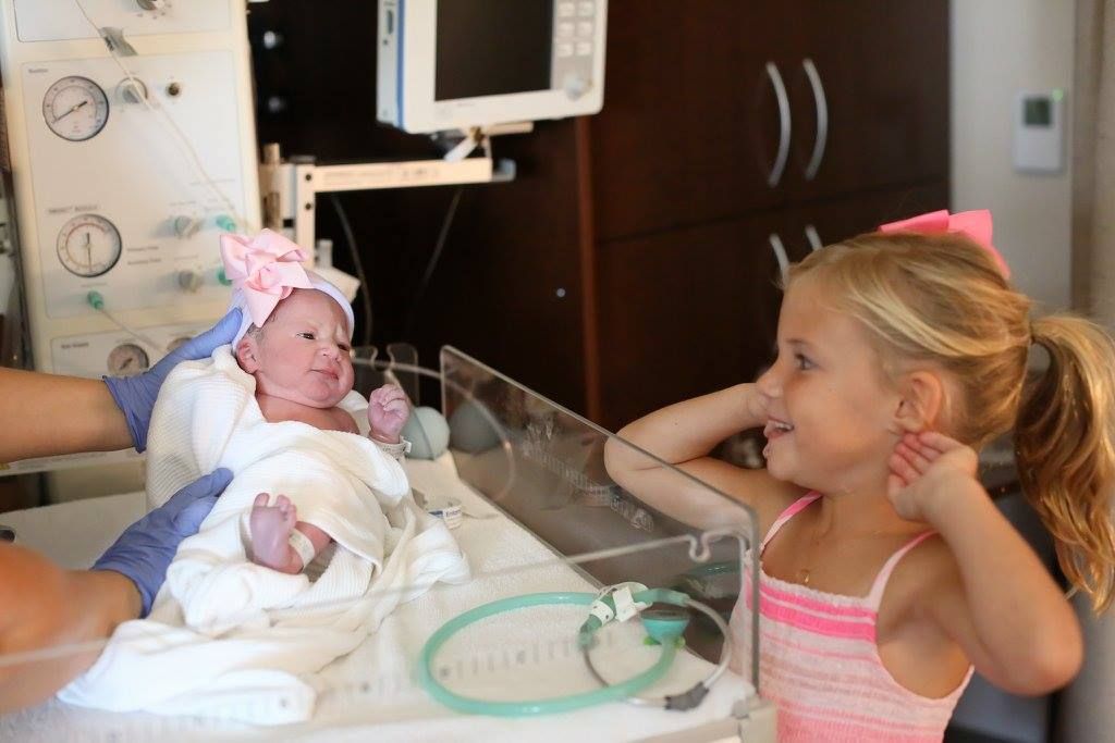 Hospital room scene showing family members gathered around newborn in bassinet.