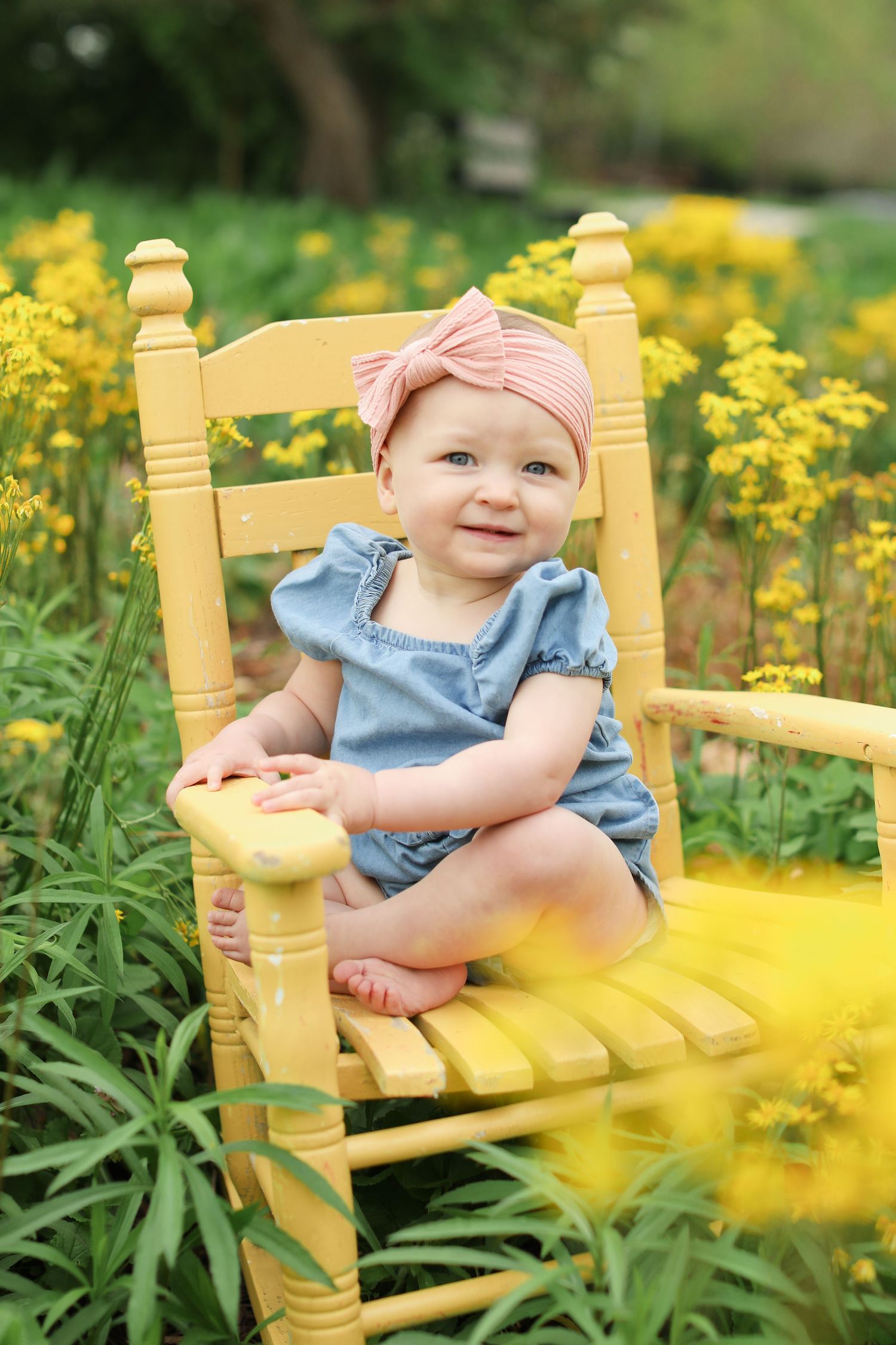 Baby in denim romper with pink headband sitting in yellow wooden chair among wildflowers.