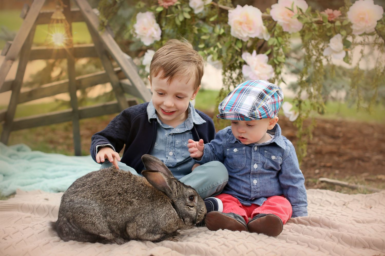 Two children sitting on blanket petting gray rabbit near rose garden.