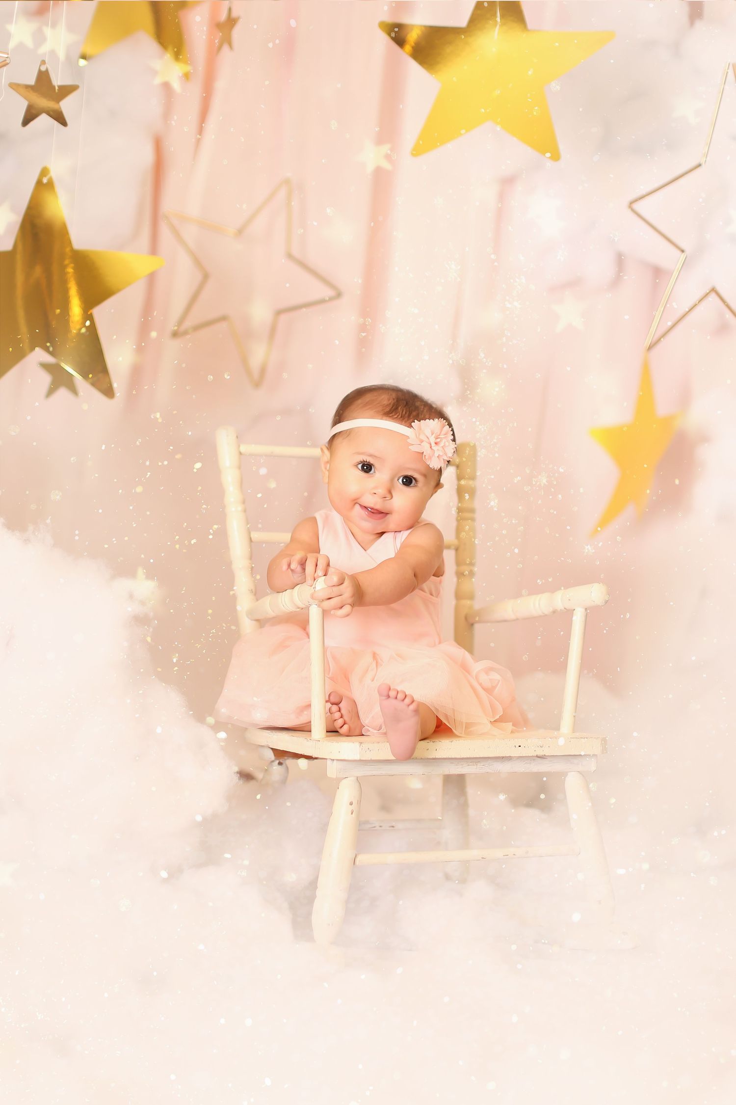 Baby in pink dress sitting on white rocking chair surrounded by golden stars and cloud backdrop.