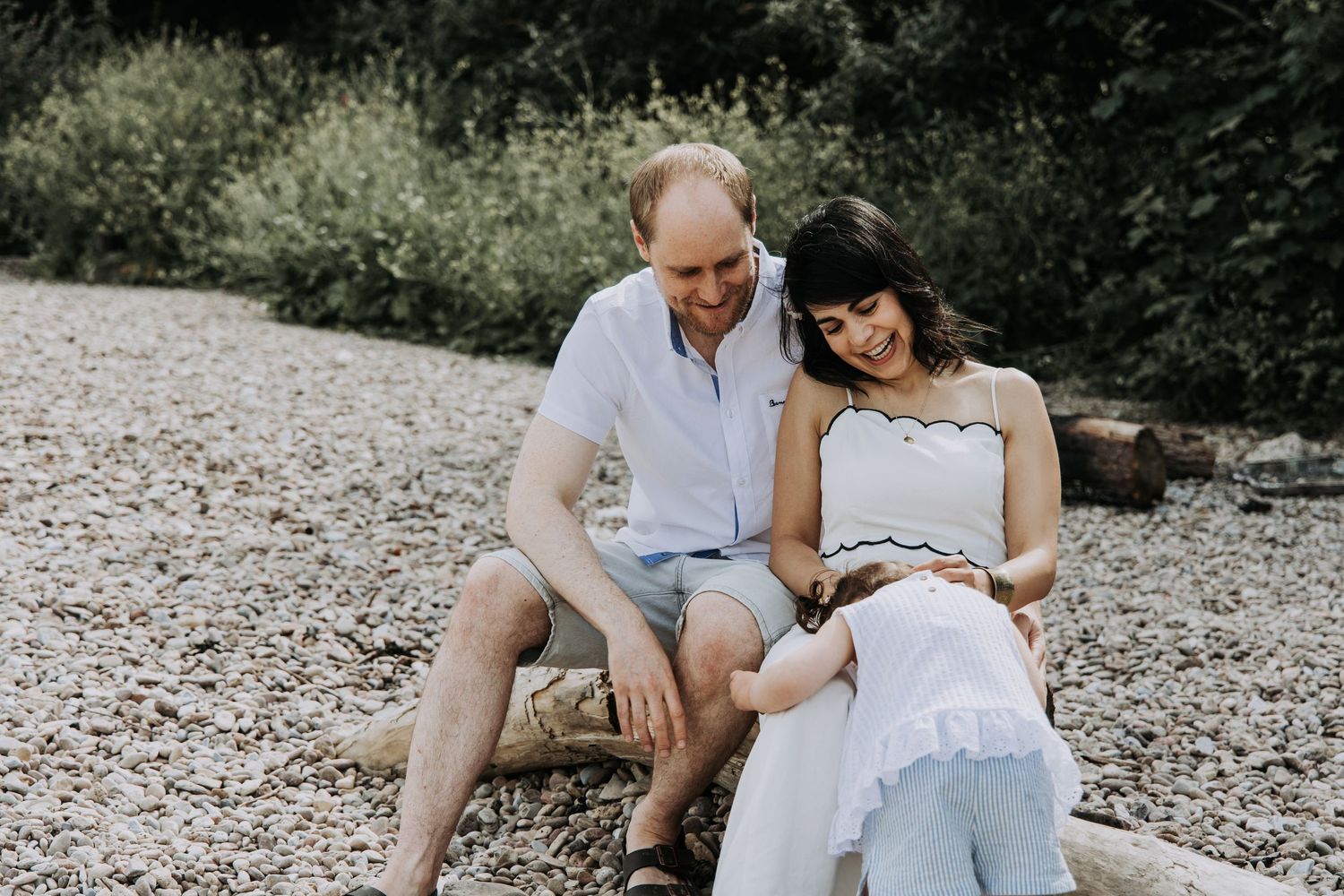 Parents sitting together while their toddler explores during a relaxed family photography session on a pebble beach.