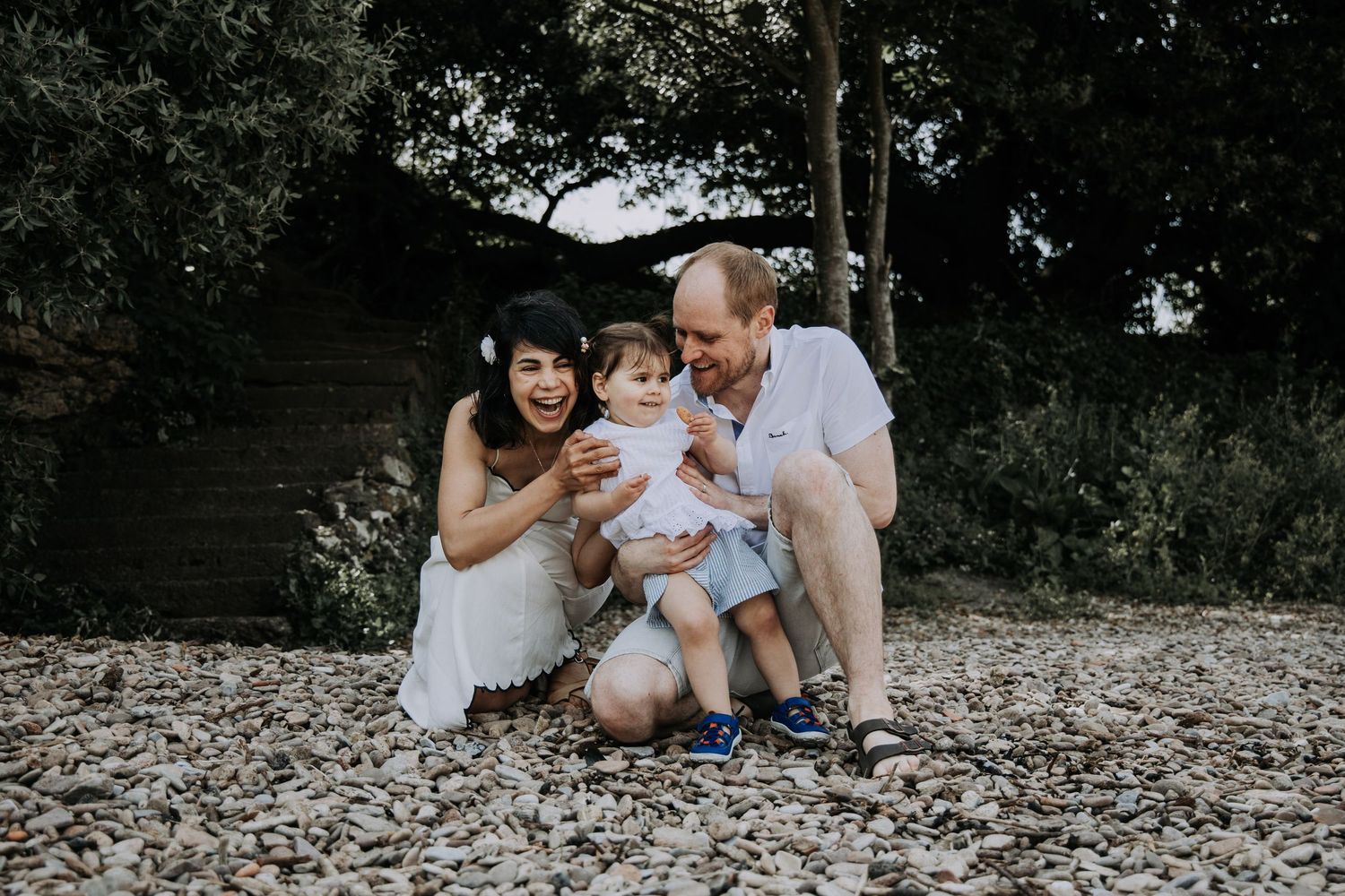 Parents laughing with their toddler on a pebble beach during a relaxed family photography session.