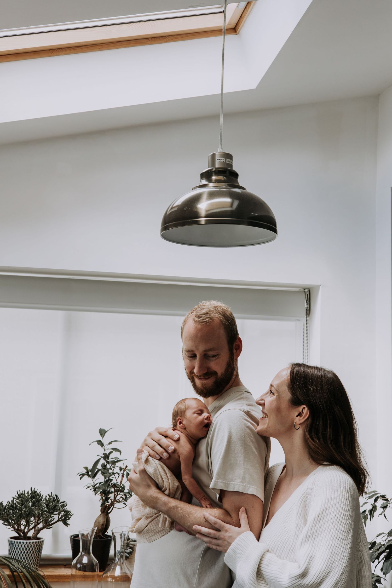 Parents smiling while holding their sleeping newborn baby in a bright, naturally lit home during a lifestyle newborn photoshoot.