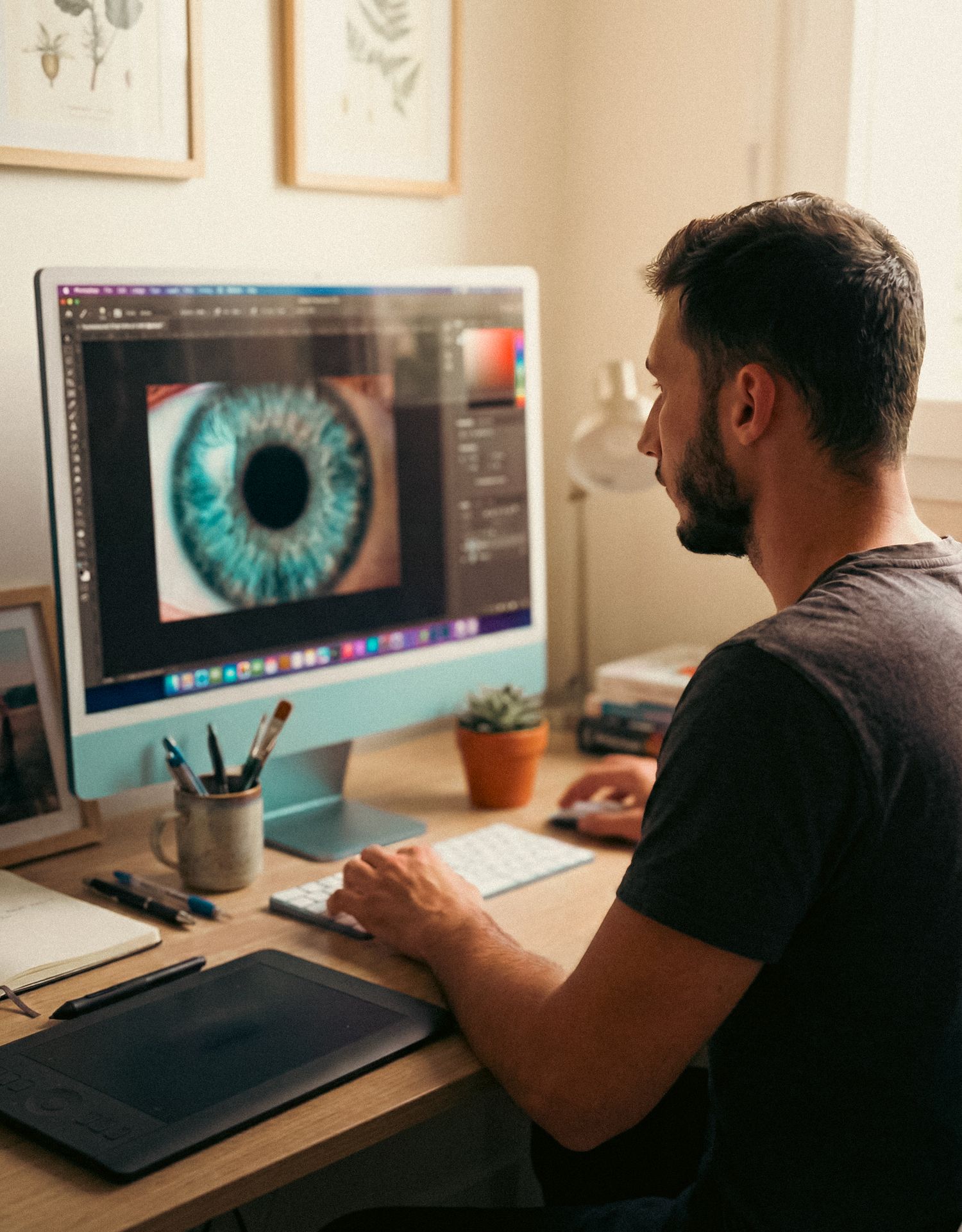 Designer working at desk with close-up views of digital artwork showing detailed eye illustrations on computer monitors.