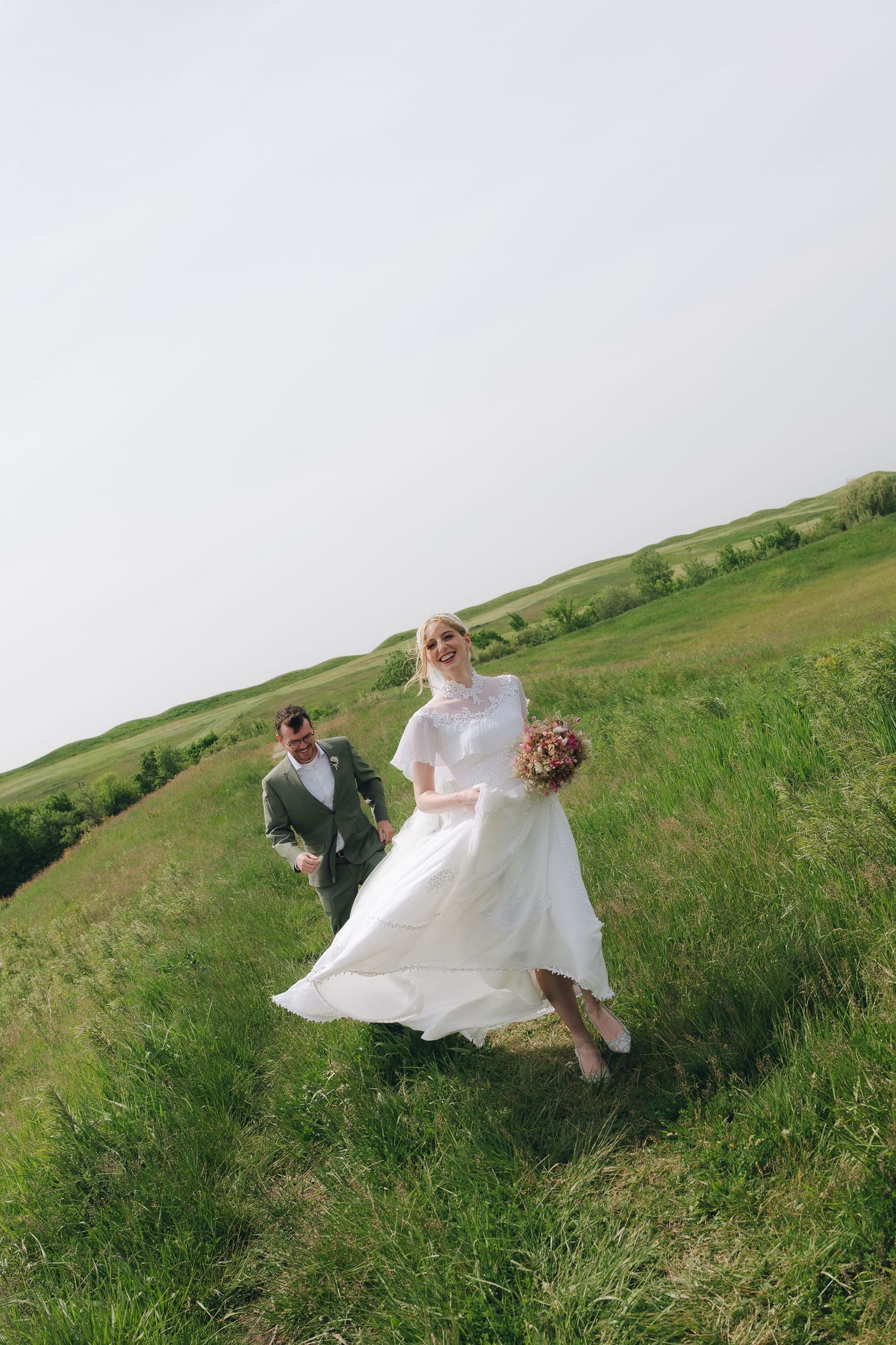 Joyful bride in white dress and groom in gray suit running through lush green meadow on their wedding day.