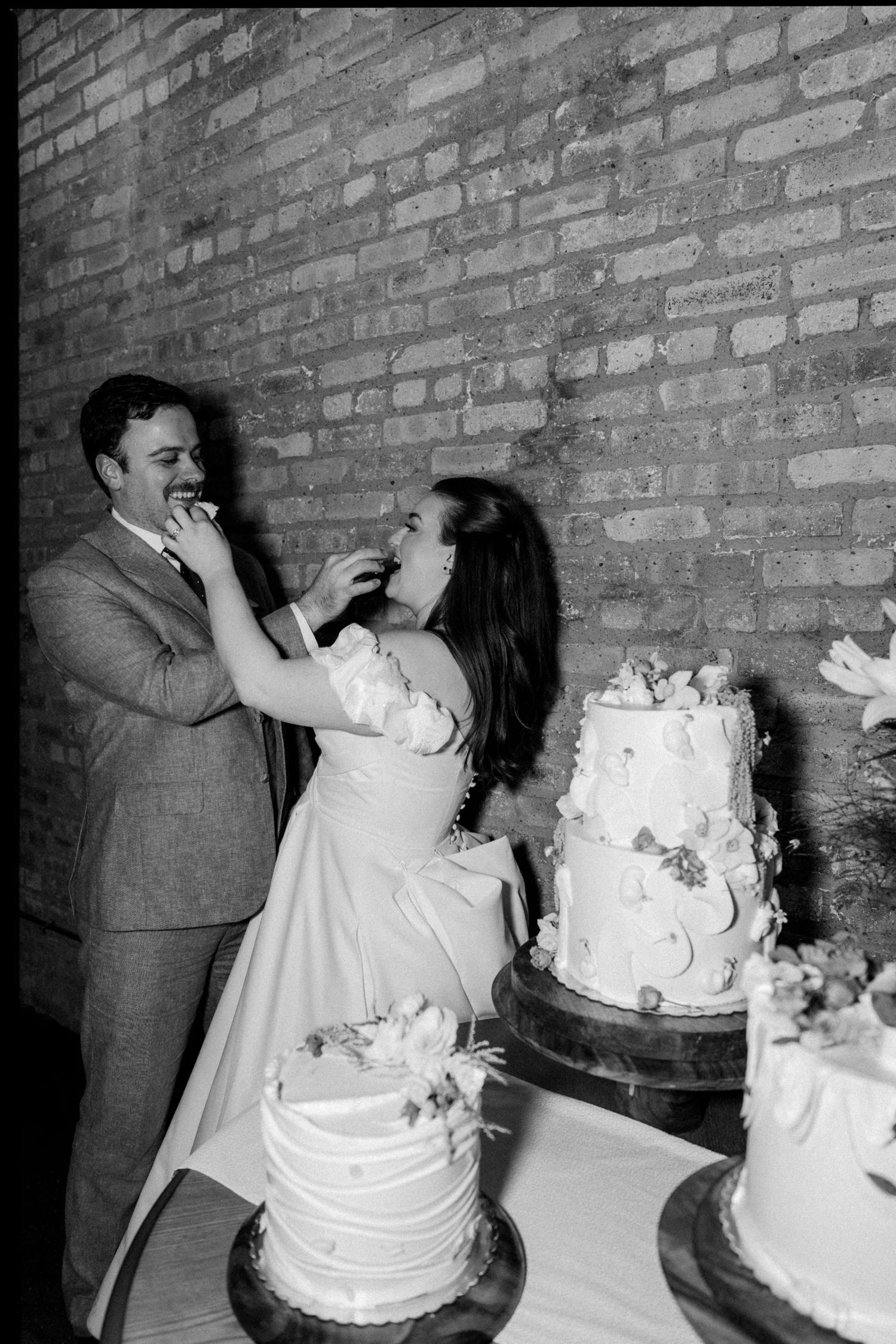 Black and white photo of bride and groom feeding each other wedding cake, laughing in front of brick wall.