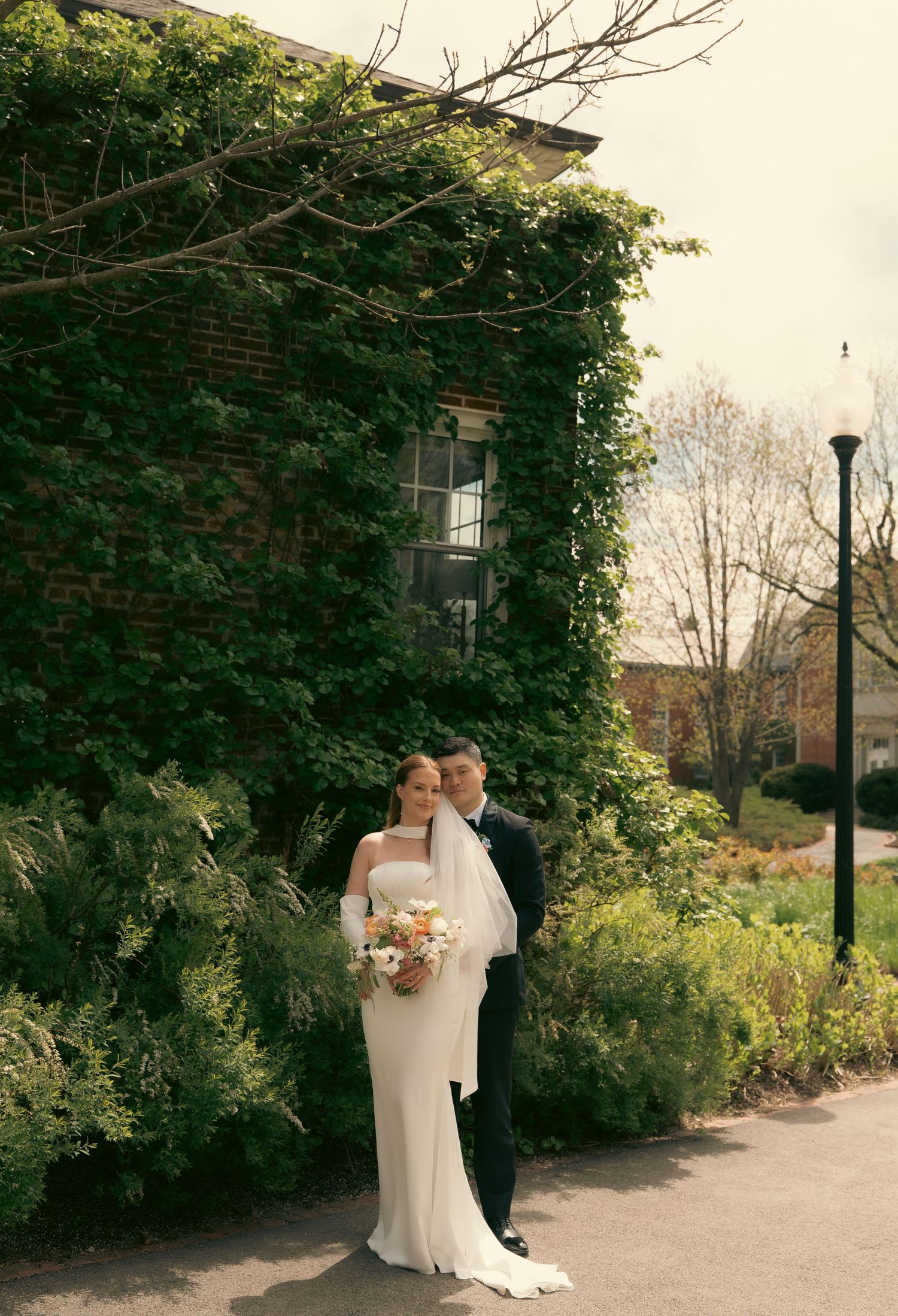Bride and groom pose together in front of an ivy-covered brick building, bride holding a colorful bouquet.