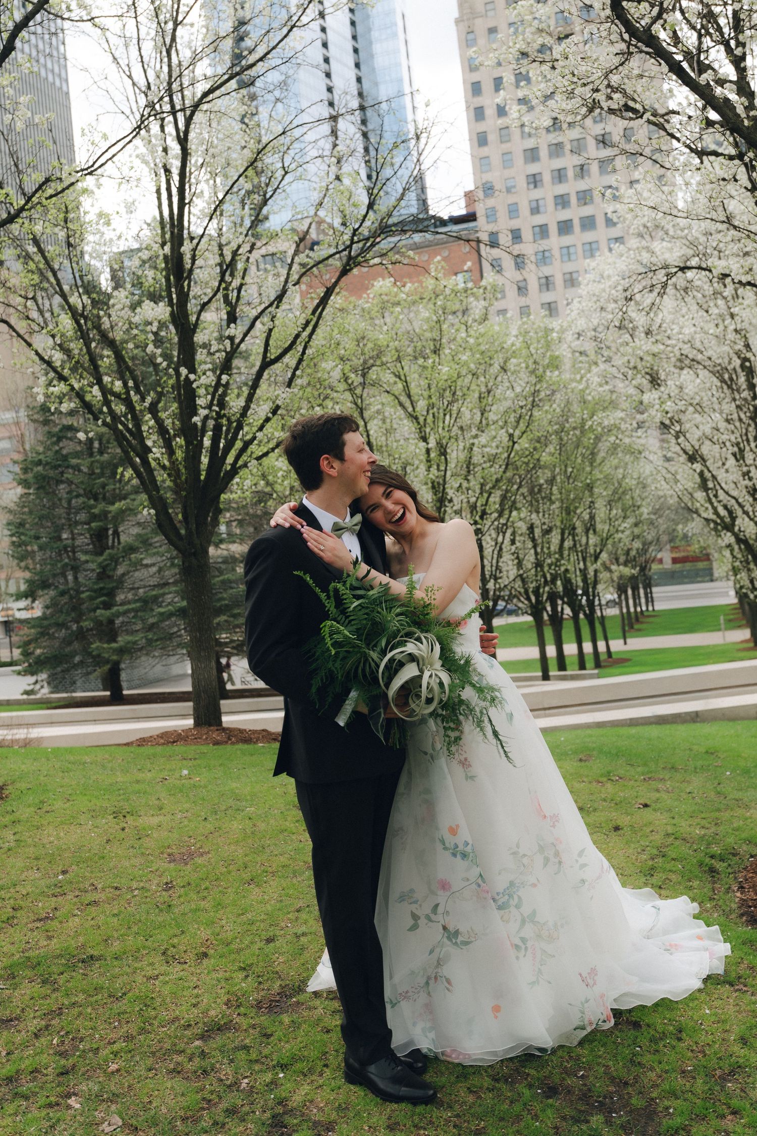 A bride and groom embrace in a park with blooming trees and city skyline, bride holding a lush green bouquet.