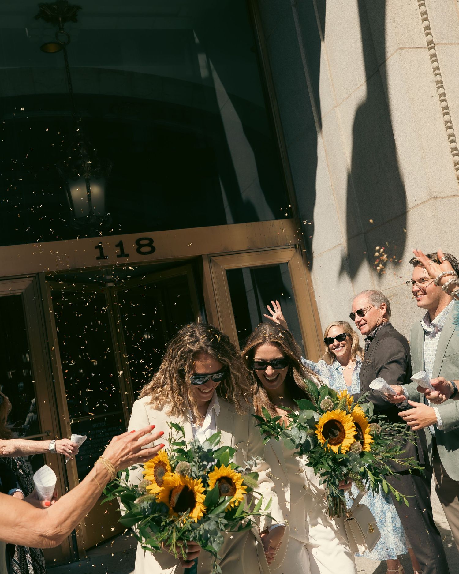 Wedding guests toss confetti in celebration as sunlight streams through a covered outdoor venue with sunflowers.