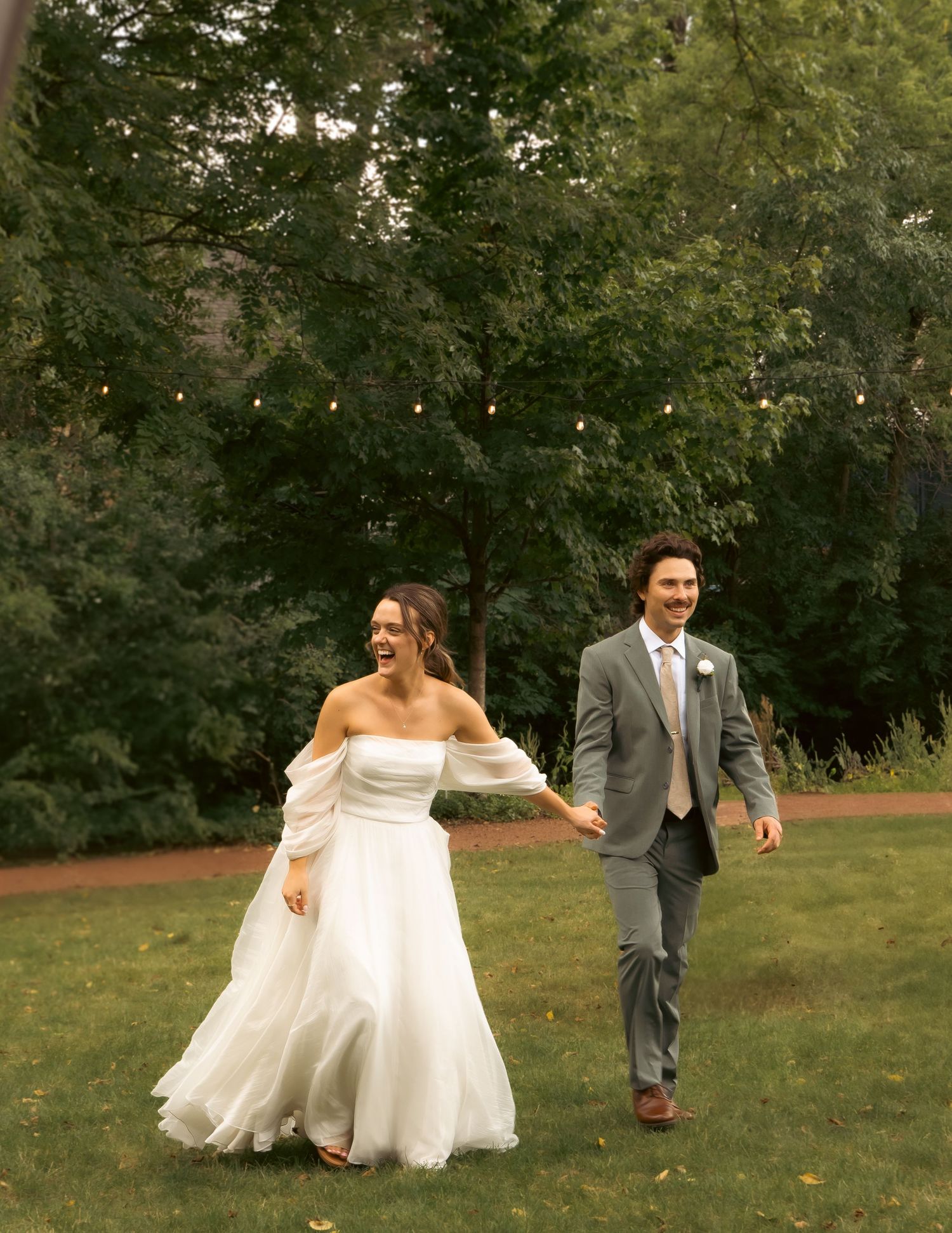 A couple in wedding attire laughs while walking together on a lawn with string lights and trees in the background.