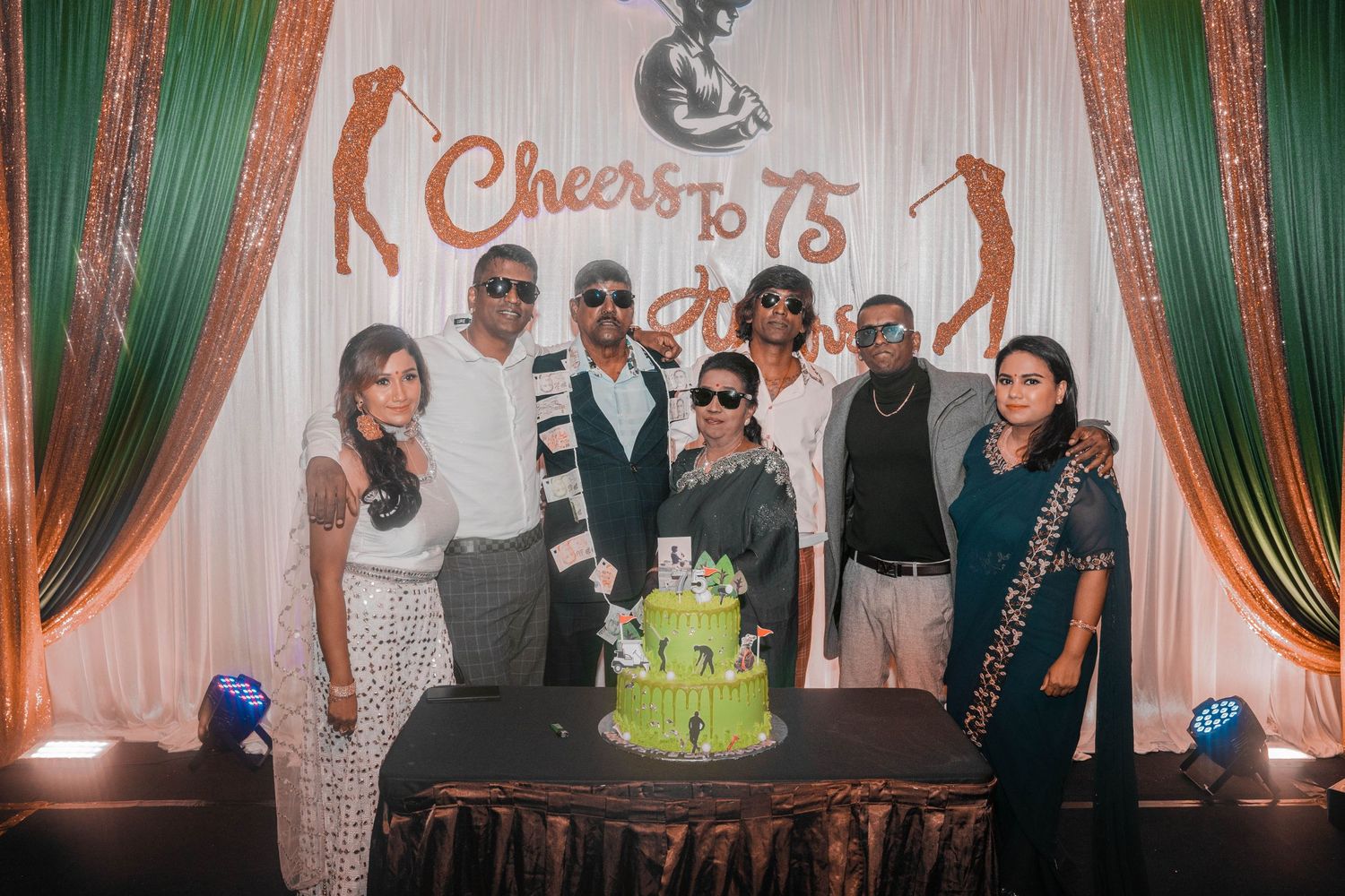 Group celebration event with green decor and a custom cake displaying 'Cheers to 75'.