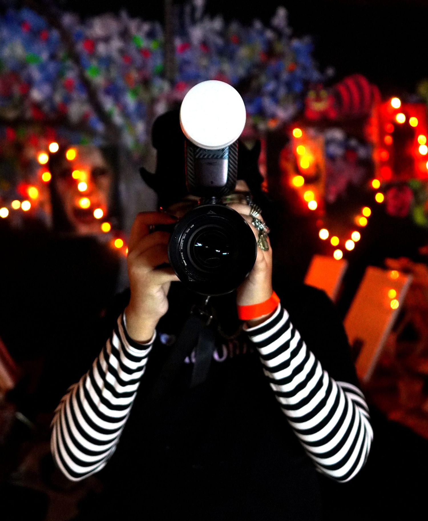 A photographer with striped sleeves holds a camera with a bright round flash in a dark setting with colorful lights in background.