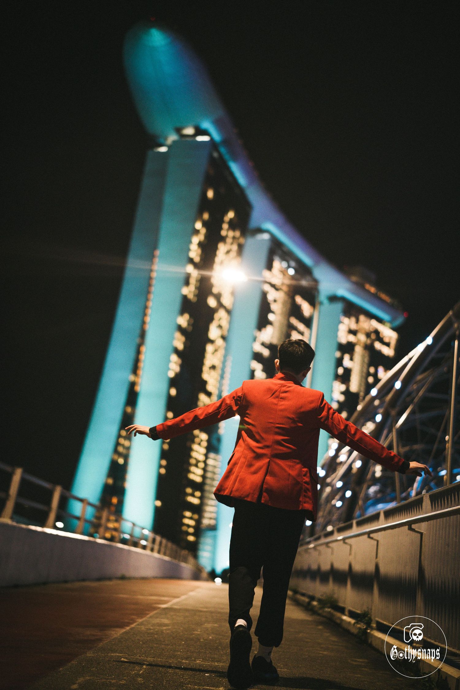 Person in red jacket stands with outstretched arms on bridge walkway with illuminated Marina Bay Sands towers in Singapore.