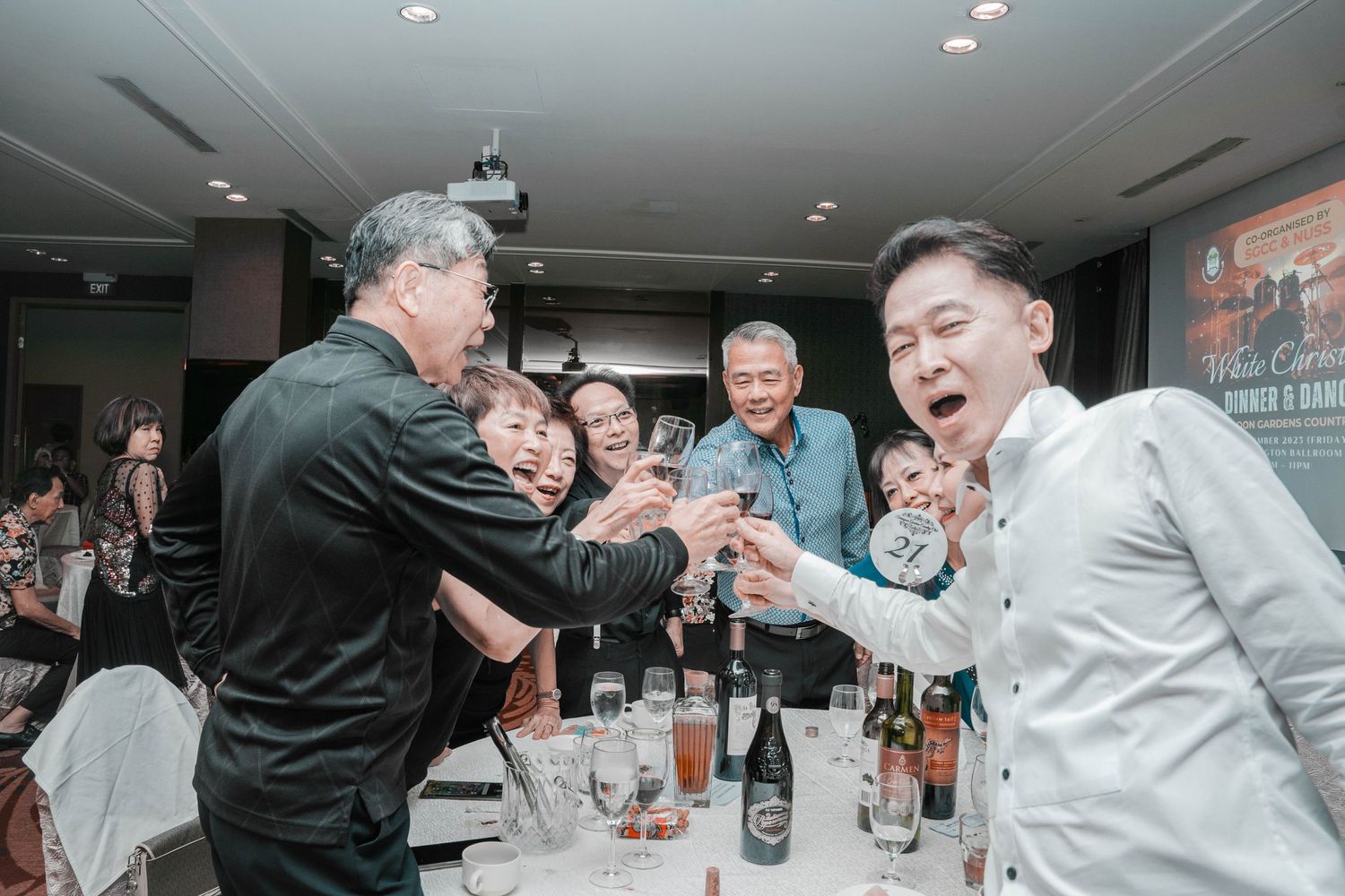 Lively dinner party scene with guests laughing and toasting at a restaurant table.