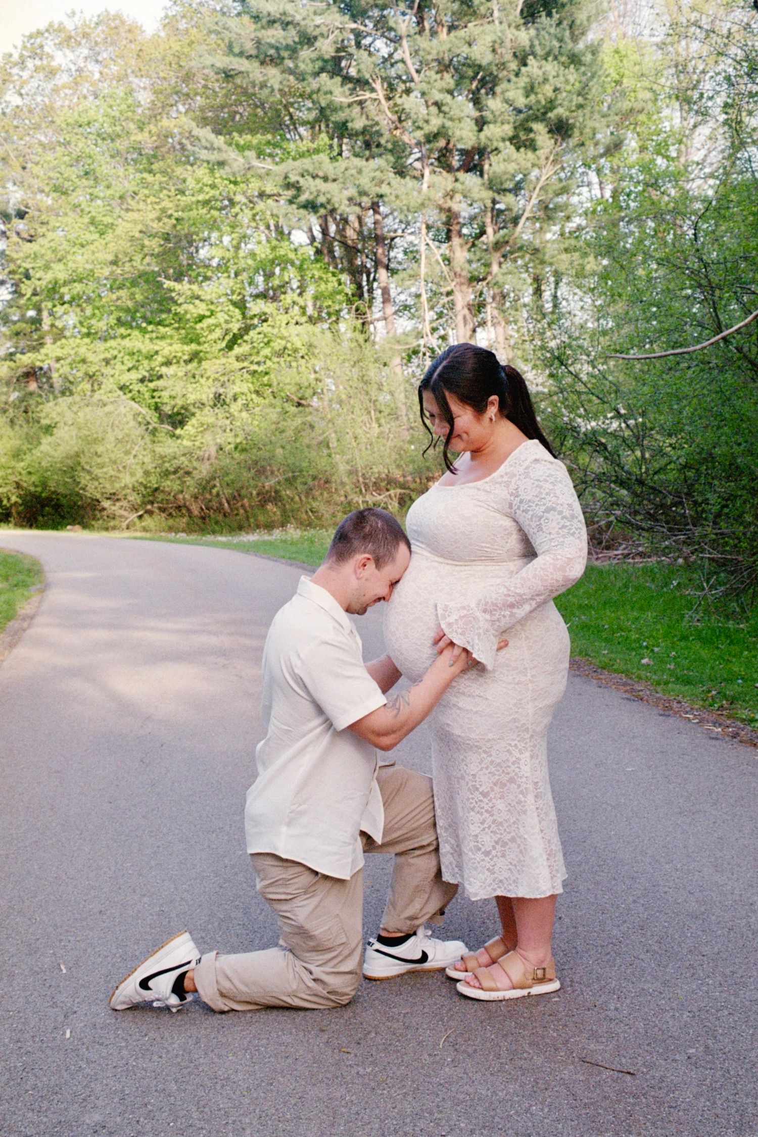 A man kneels before his pregnant partner on a tree-lined path, gently touching her baby bump during a maternity photo session.