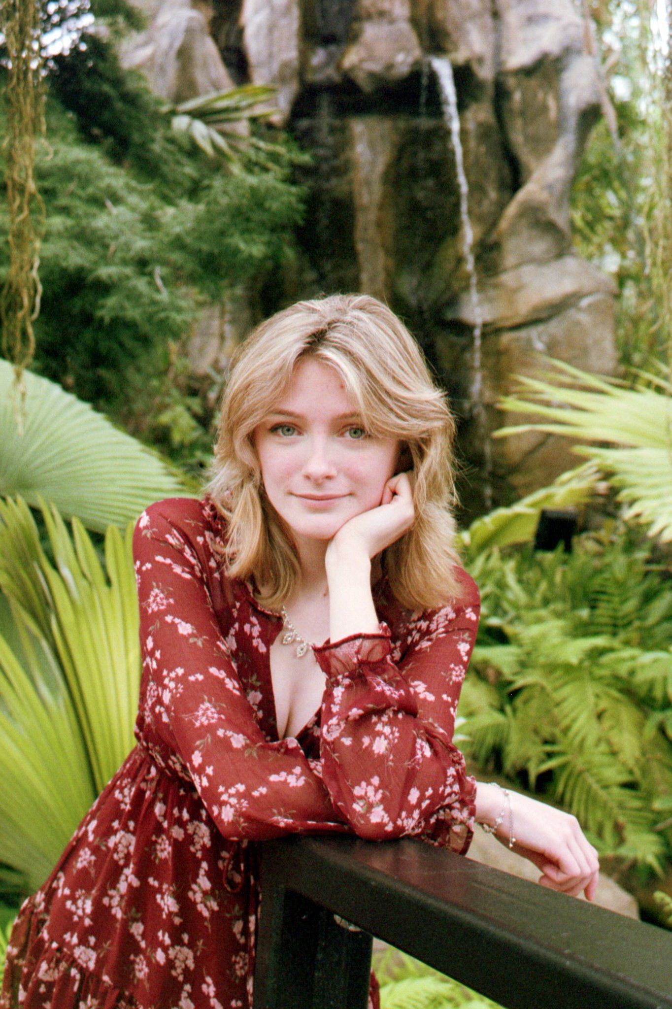 Young woman in red floral dress leaning on wooden railing in lush tropical garden with waterfall.