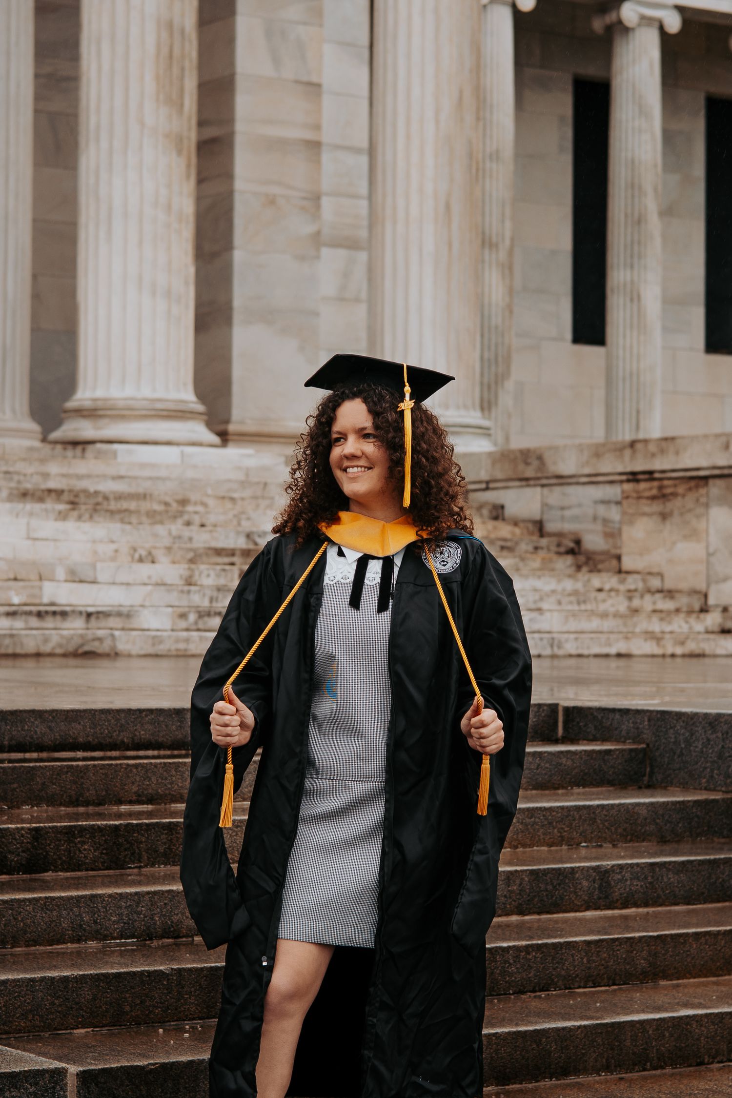 A graduate in cap and gown poses on steps of a neoclassical building.