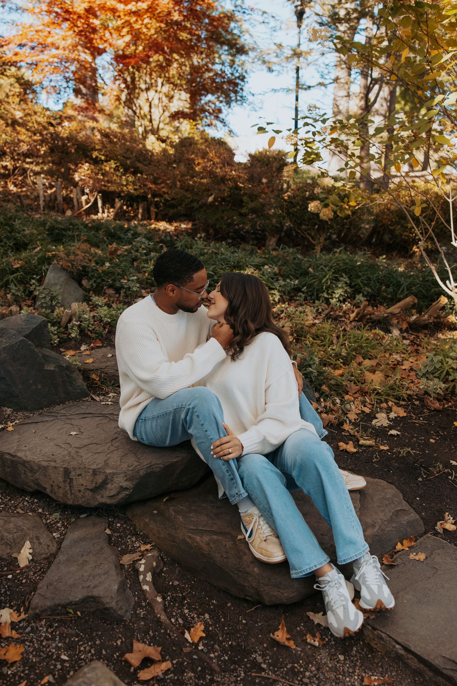 Couple in casual wear sharing romantic moment on rocks surrounded by fall foliage.