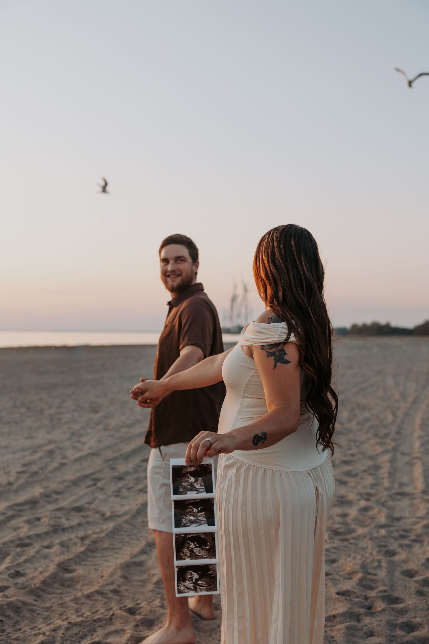 Couple on beach at sunset holding ultrasound photos, woman in white dress with tattoos, man in brown shirt smiling.