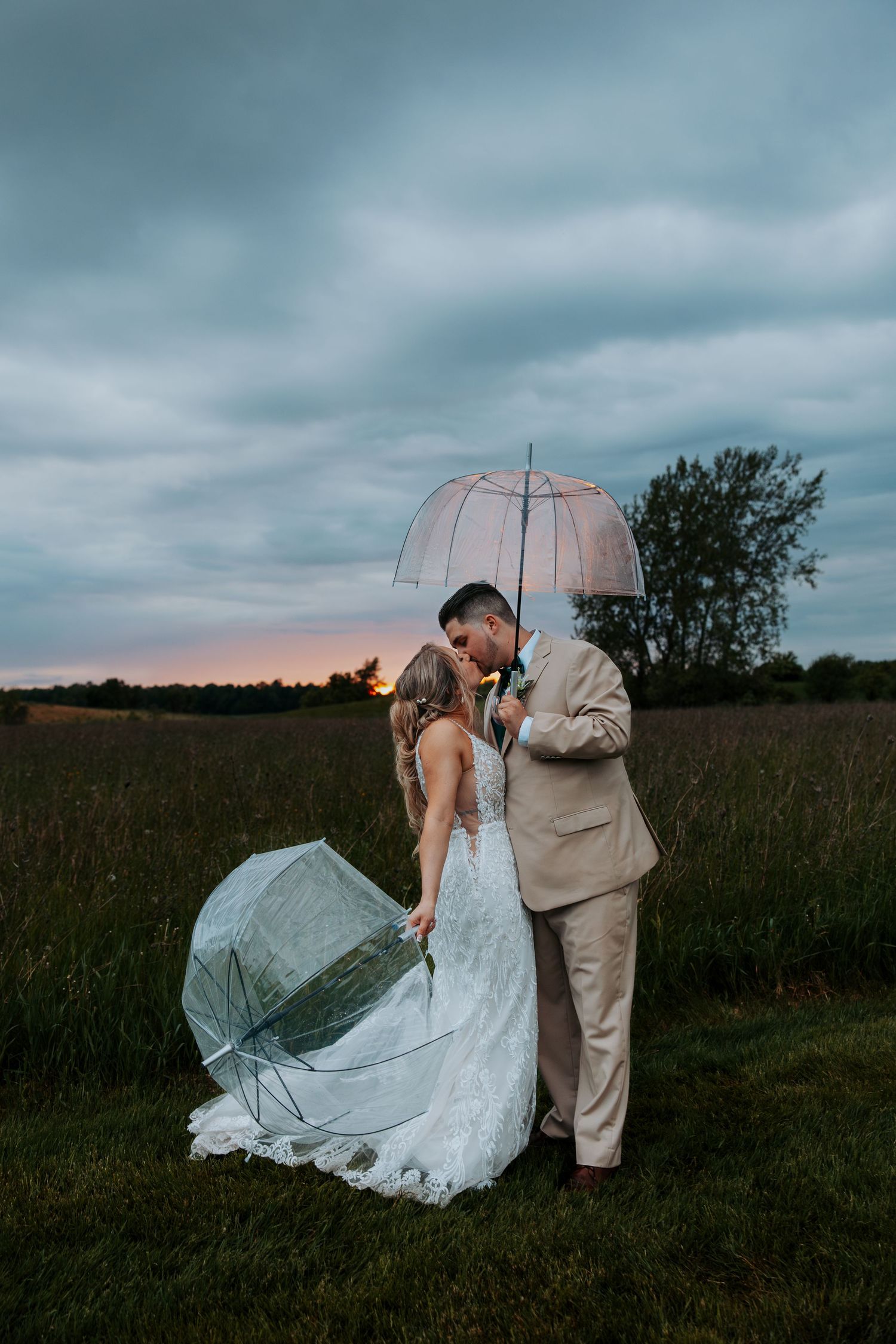 Couple in wedding attire walking through field during moody sunset with flowing dress.
