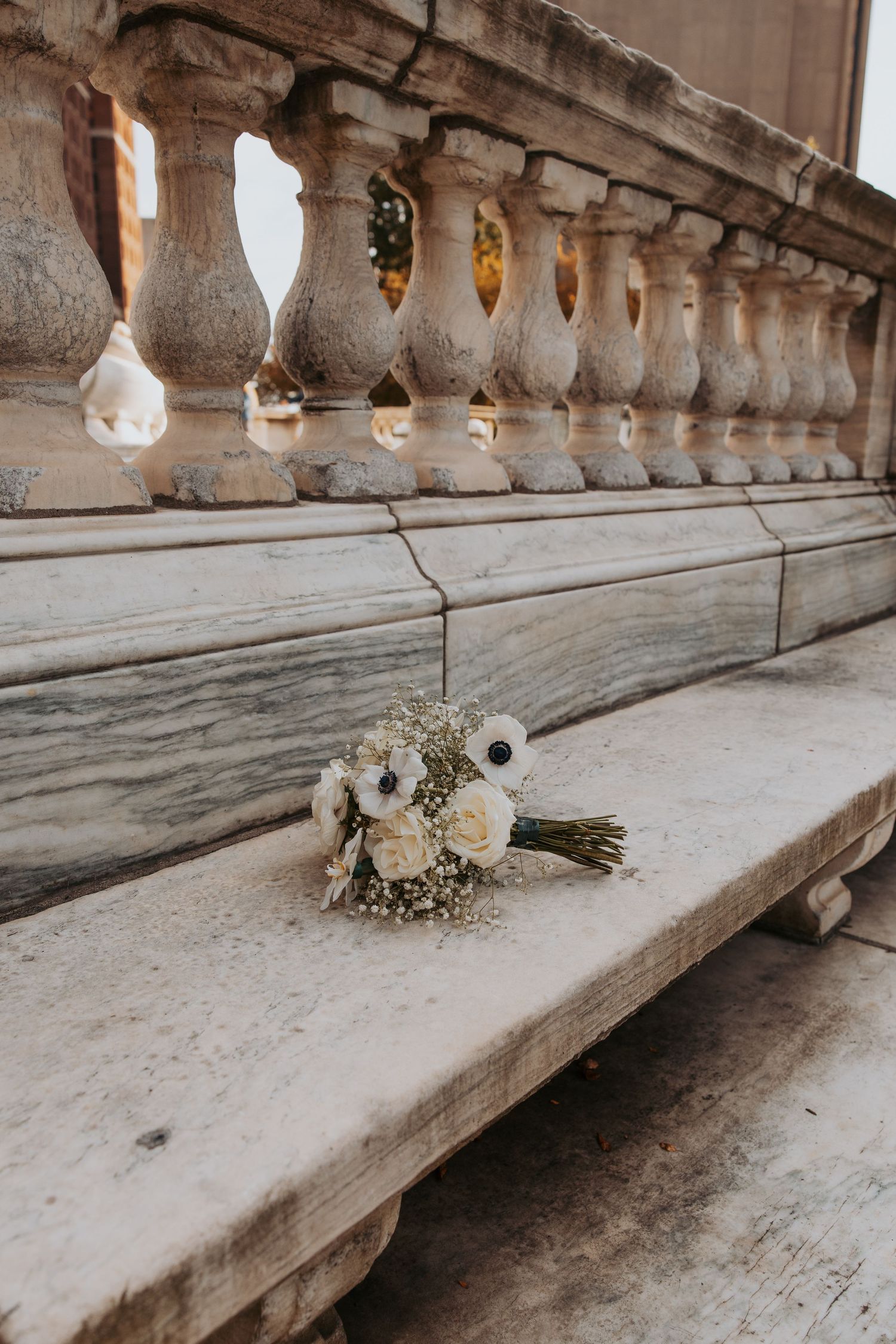 White wedding bouquet with anemones and baby's breath resting on marble steps beside ornate stone balustrade.