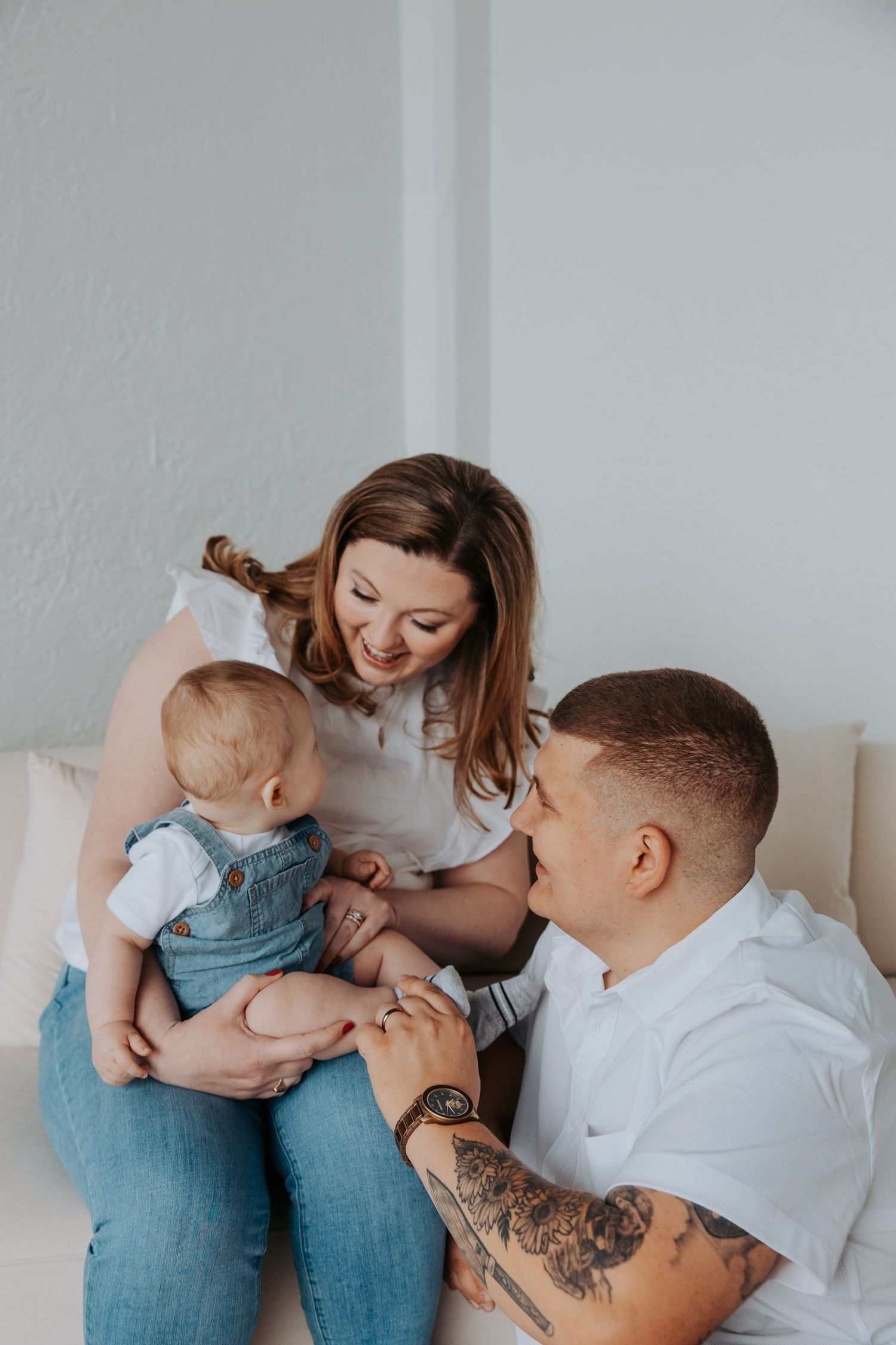 Happy young parents smiling at their baby dressed in denim overalls while sitting together on a white couch.