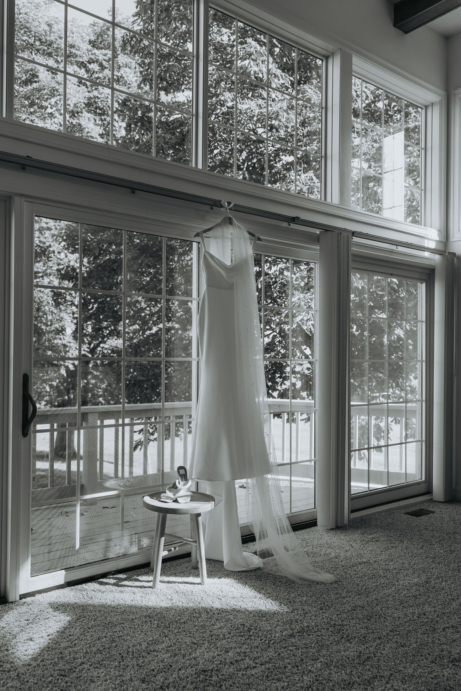 Black and white photo of a wedding dress hanging by large windows with a small stool and veil below.